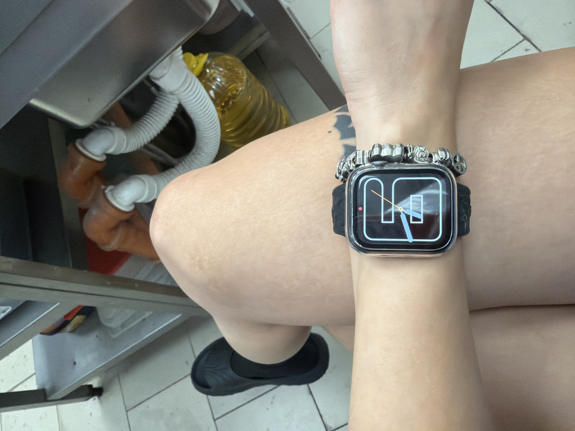 The image shows a close-up of a person's arm wearing a smartwatch and a charm bracelet. The smartwatch displays a minimalist analog clock face with two thin hands. The arm is bare and has a tattoo on the upper part. The person is sitting, with their legs visible in the foreground. They are wearing black socks and black slip-on shoes.
In the background, there is plumbing from what appears to be a sink, with white flexible pipes and orange plastic pipes. A large bottle of yellow liquid, possibly cooking oil, is also visible. The setting appears to be indoors, likely a kitchen or a utility area, given the presence of plumbing and the tiled floor.
The lighting suggests it is daytime. There are no explicit emotional cues, but the overall scene is mundane and focuses on the person's accessories and immediate surroundings. No visible text is present in the image. The provided location context is Plovdiv, Bulgaria.
