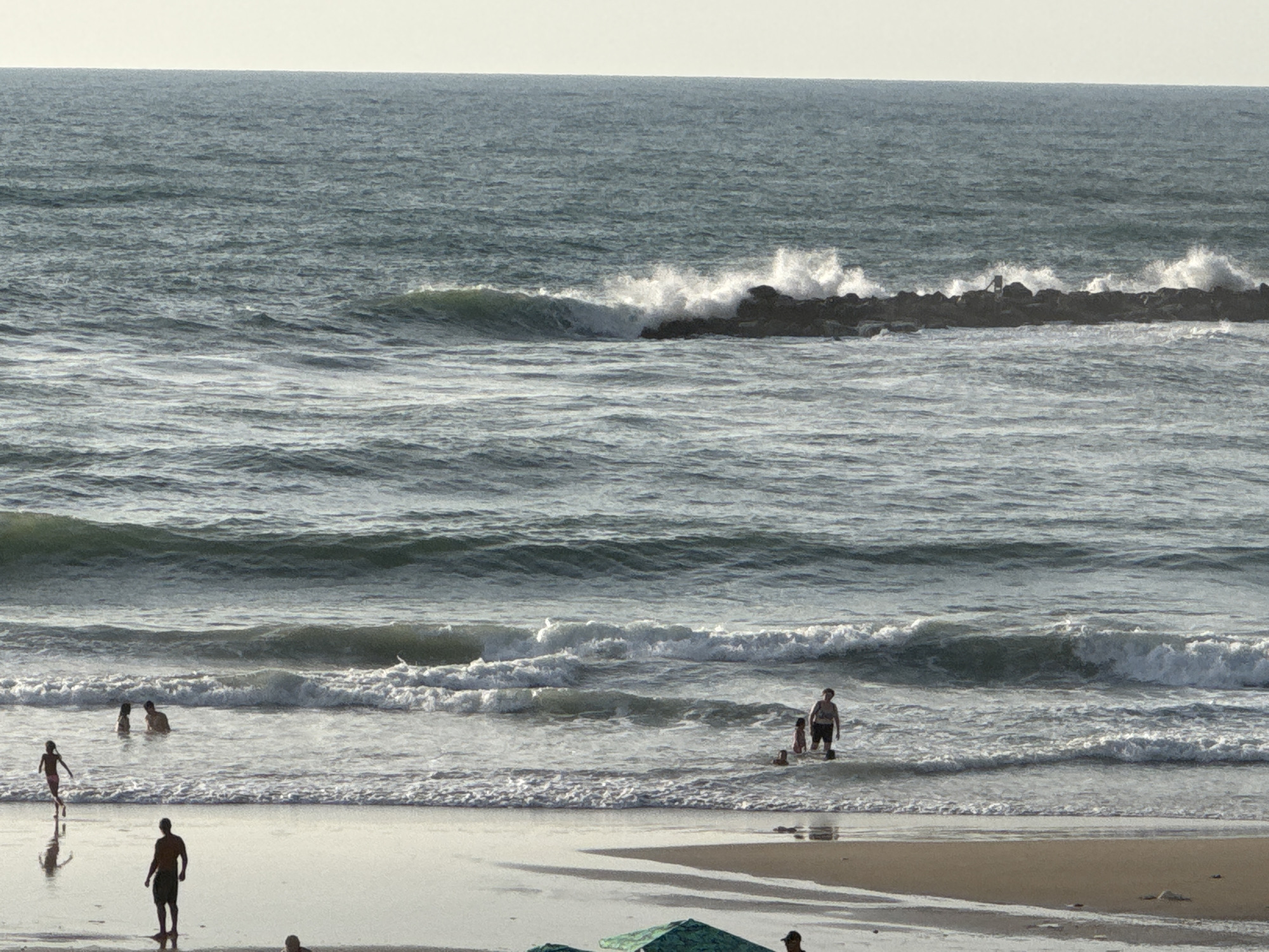 The image depicts a beach scene in Netanya, Israel, with the Mediterranean Sea in the background and a sandy shore in the foreground. Several people are visible in the water and on the beach.
In the water, closer to the shore, a woman wearing a dark swimsuit stands with a young child next to her. Further out, a group of at least three people are partially submerged in the water, appearing to be swimming or wading. A lone man, wearing dark shorts, stands in the shallow water near the shore. To the left, a child wearing a pink swimsuit runs along the water's edge.
On the beach, a man wearing dark shorts is walking away from the viewer towards the water. In the far background, a rocky breakwater stretches across the sea, with waves crashing against it, creating white foam. The sky is a pale, overcast color, and the water is a deep blue-green, reflecting the muted light. The sand is wet and reflective near the waterline, with dry sand further up the beach. Two green beach umbrellas are visible at the bottom of the frame, partially obscuring the view of the sand.