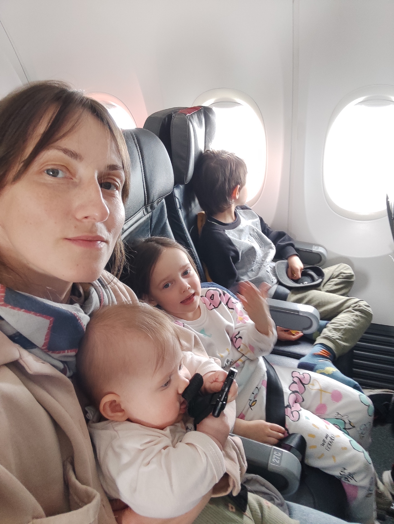 A medium shot depicts a woman with three children on an airplane. The woman is in the foreground, facing forward, with a baby in her arms. The baby is holding a black object in its hands. Behind the woman and baby is a young girl, also facing forward and wearing pajamas. In the background, seated by the window, is a young boy looking out the window. The airplane's interior is visible, including the seats and windows. The location in the city of Chaikovsky, Russia, is not determinable from the image.
