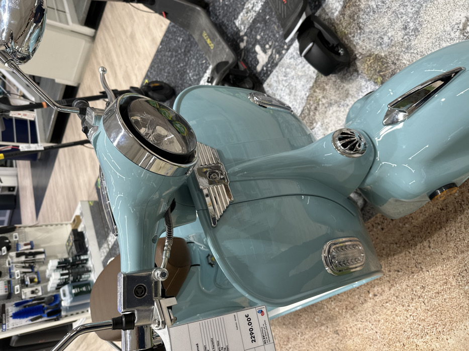 The image displays a light blue (aqua/mint green) retro-style scooter, likely on display in an indoor retail showroom or a large store in Metamorfosi, Greece. The scooter is positioned at an angle, highlighting its front and right side. It features a classic design with a prominent round headlight encased in a chrome bezel, chrome handlebars, and a chrome left-side mirror. Additional chrome accents include a decorative vertical grille on the front fairing and small circular vents near the handlebars. A segment of a brown seat is also visible.
A white price tag is attached to the scooter, prominently displaying "2290.00€". Below the price, Greek text is visible, including "ΚΛΑΣΣΙΚΟ" (Classic), "Αγοράστε τώρα" (Buy now), and "με 12 άτοκες δόσεις" (with 12 interest-free installments). The tag also identifies the retailer as "Δ. ΚΩΤΣΟΒΟΛΟΣ ΑΕΒΕ" (Kotsovolos), a known Greek electronics and home appliances chain. The left-side mirror bears the inscription "E4 000130".
In the background, an electric scooter, possibly a Segway-Ninebot model with "NINEBOT" visible on its footboard, is partially seen. To the left of the scooter, shelves stocked with various products, potentially automotive accessories or cleaning supplies, line the wall, consistent with a retail environment. The flooring includes a textured grey section and a lighter, possibly wood-look or tiled area. No people are present, and no specific activity is taking place other than the display of items for sale under artificial indoor lighting.