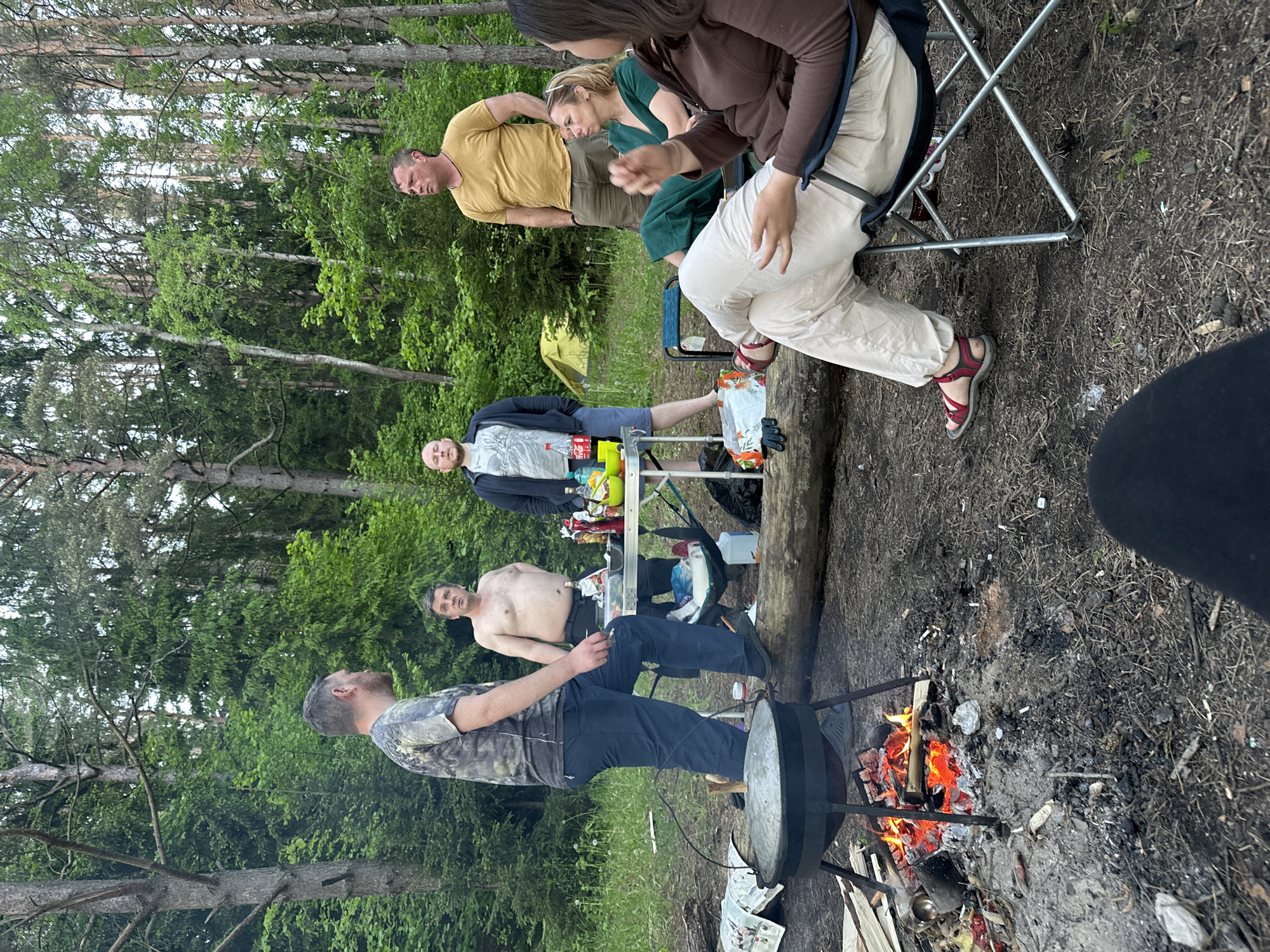 Location: Outdoors in a wooded area, likely near Юзуфоўскі сельскі Савет, Belarus.
Scene: Several people are gathered around a campfire, seemingly engaged in a camping or picnic event. A man in a camouflage shirt is positioned near the fire, possibly cooking in a large pot suspended over the flames. Another man is standing shirtless near a folding table laden with food and drinks. A third man lies on the ground near the table. A woman in a green dress is standing, and a man in a yellow shirt stands beside her. A person with brown hair is sitting in a camping chair.
Objects: Campfire, large cooking pot, folding table, food, drinks, camping chair, trees, wood.
Actions: Cooking, eating, socializing.
