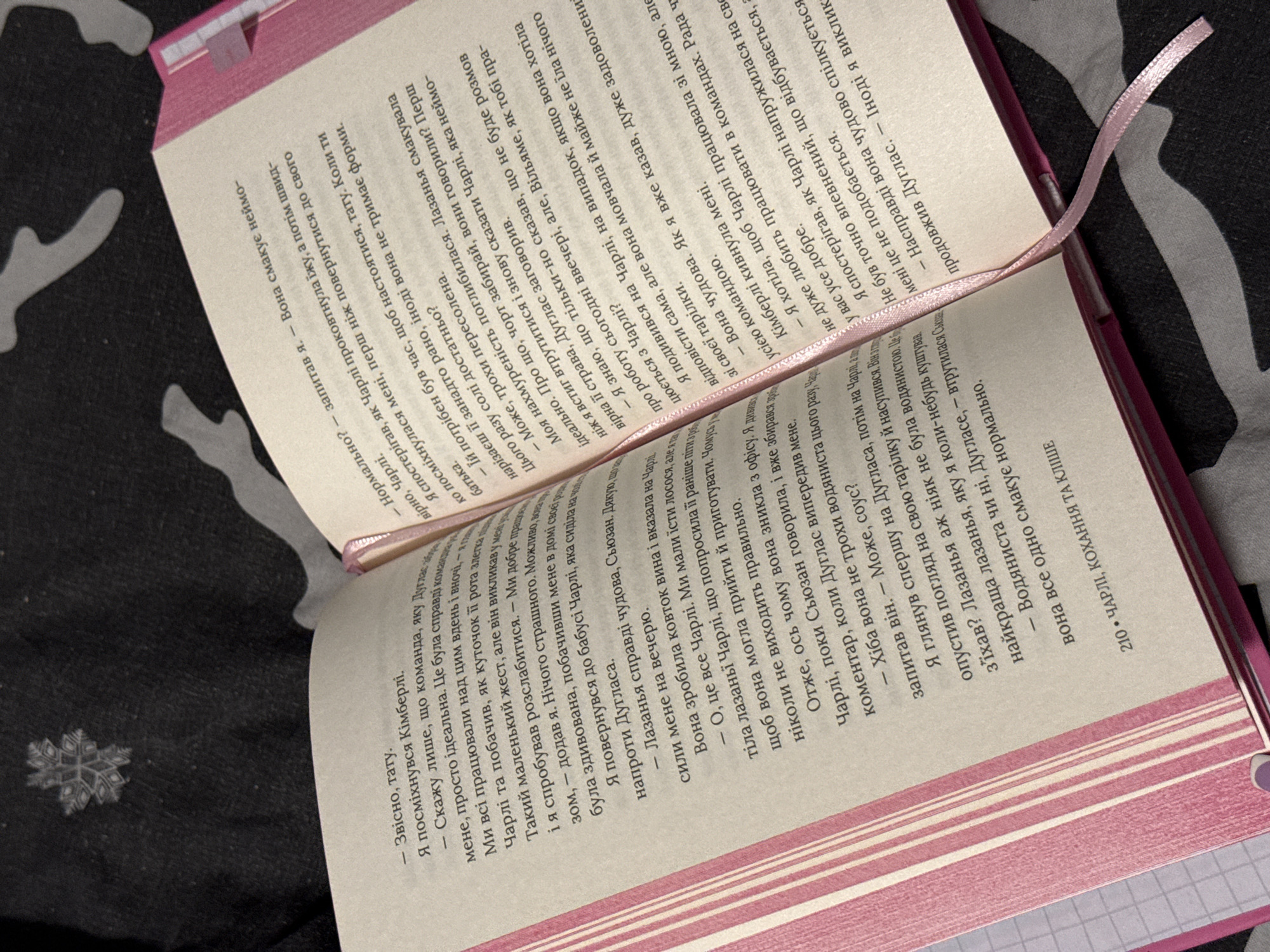 The image shows an open book with pink pages and a pink ribbon bookmark. The text is in Ukrainian. The book is lying on a dark surface with a subtle pattern. The lighting suggests an indoor setting. There are no people or any discernible signs of the city of Łódź, Poland, or any other specific location. The scene is a close-up of the book and its immediate surroundings.