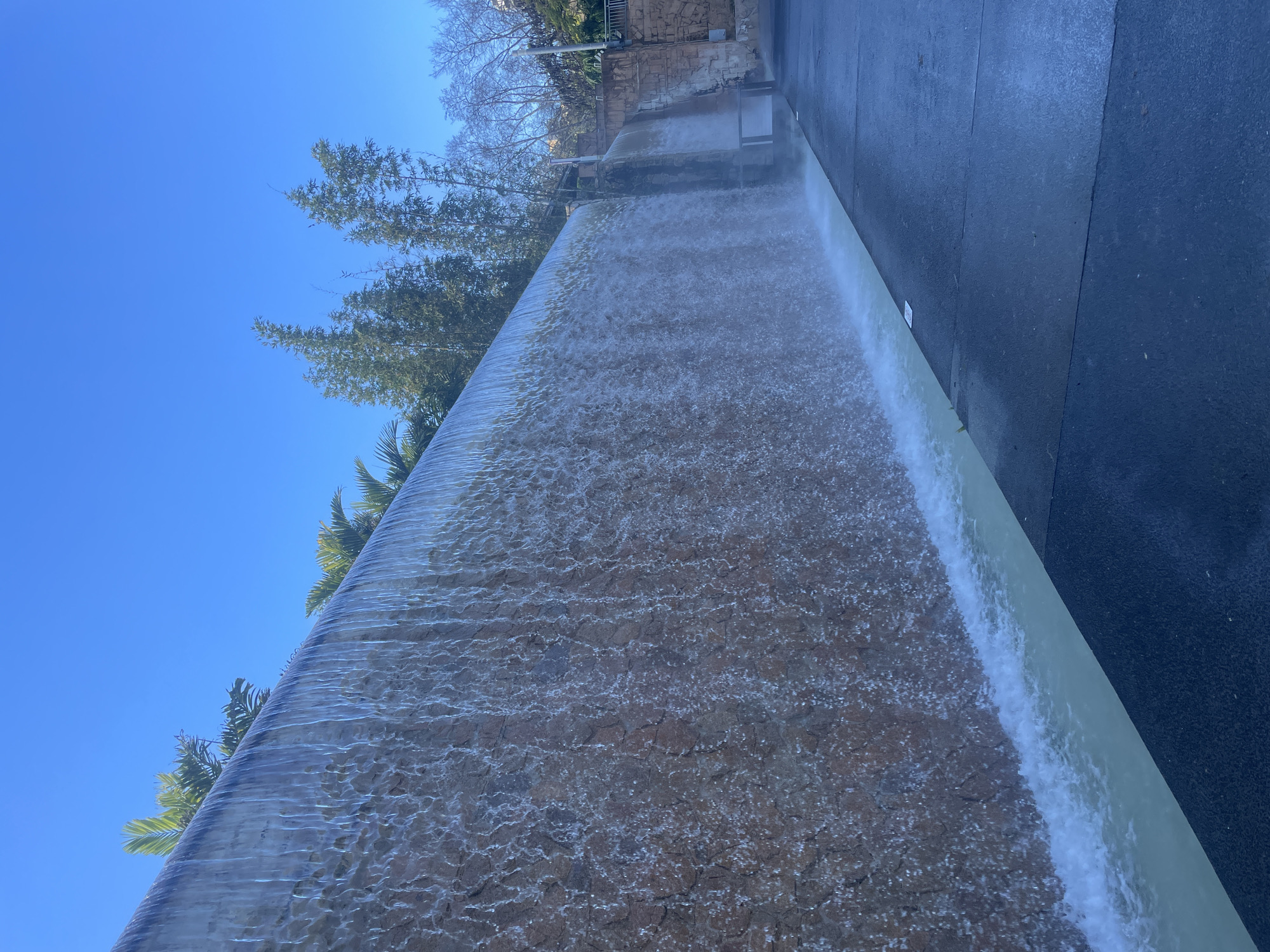 The image depicts the iconic Waterfall in South Bank Parklands, Brisbane, Australia. A wide, artificial waterfall cascades down a textured stone wall into a shallow, light-colored pool at its base. The water is clear and creates a shimmering curtain as it falls.
To the left of the waterfall, lush green palm fronds are visible peeking over the top edge, suggesting a tropical or sub-tropical climate. Behind the main waterfall, further to the right, another, smaller waterfall can be seen, flowing down a more traditionally constructed stone wall.
The foreground is dominated by a dark, paved pathway that runs alongside the pool. The pathway appears wet in places, likely from mist or spray from the waterfall. The sky above is a vibrant, clear blue, indicating a sunny day.
In the background, beyond the waterfalls, there are tall, slender bamboo trees and bare-branched deciduous trees, contrasting with the evergreen palms. Further in the distance, there are hints of buildings or structures, and a metal railing is visible, suggesting a public area or park. The overall scene is one of a man-made water feature integrated into a park setting, bathed in bright daylight. There are no people visible in the image.