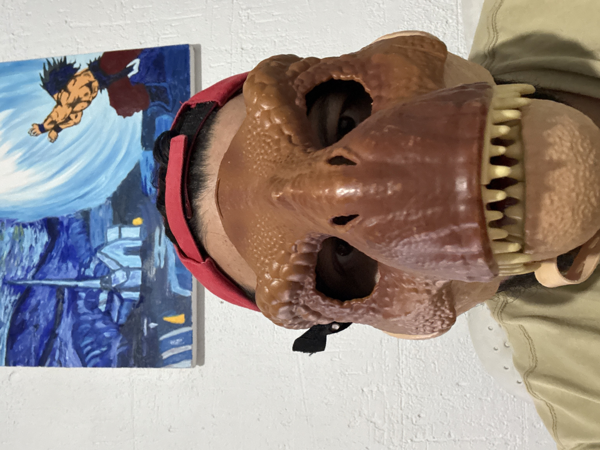 A medium shot photograph, taken from a frontal perspective, shows a person wearing a brown dinosaur mask with yellow teeth and a red cap. The person is also wearing a light brown t-shirt. In the background, a painting hangs on a white textured wall, depicting a blue, starry night sky over a village, with a muscular anime character in the upper right corner. The painting appears to be on canvas. The scene is indoors, likely in a residential setting. No specific location in Cuernavaca, Mexico can be identified from the image.