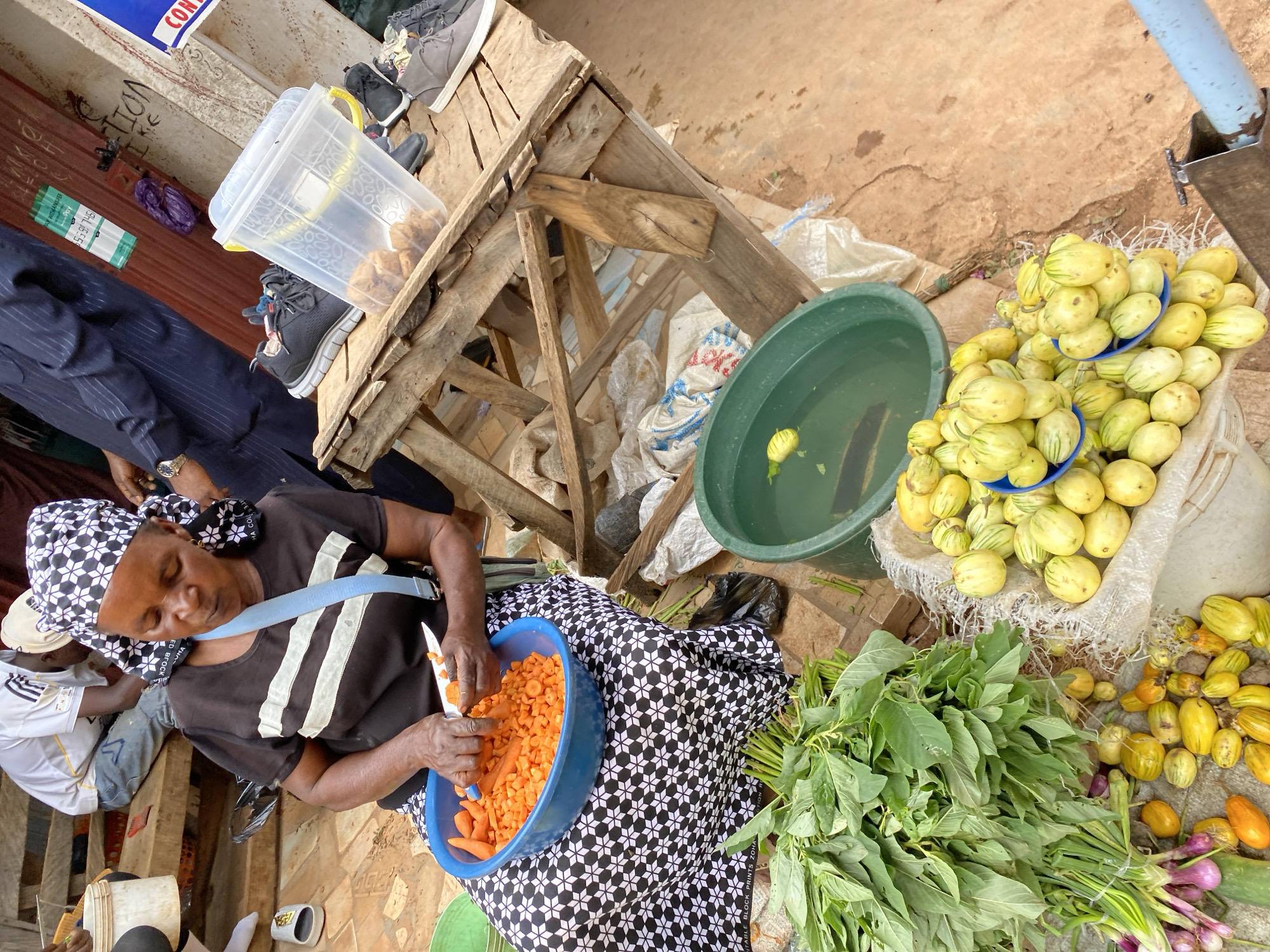 In the city of Bukuru, Nigeria, a woman with a patterned headscarf is actively preparing food at an outdoor market. She is seated, dressed in a black t-shirt with white stripes and a black and white patterned skirt, and is diligently chopping carrots into a blue bowl with a silver knife. Next to her, on a rustic wooden stand, a clear plastic container holds small, fried food items, possibly snacks. Several pairs of sneakers are visible on and around the stand. In the foreground, a generous bunch of leafy green vegetables and a pile of small, striped eggplants are displayed. A large green basin filled with water and a single yellow object, possibly a flower or piece of fruit, is situated to the right of the vegetables. The ground is a dusty, reddish-brown, characteristic of the local terrain. Other people are present in the background, some in motion, suggesting a bustling market environment. A red shuttered building with some signage is visible in the upper right portion of the image.