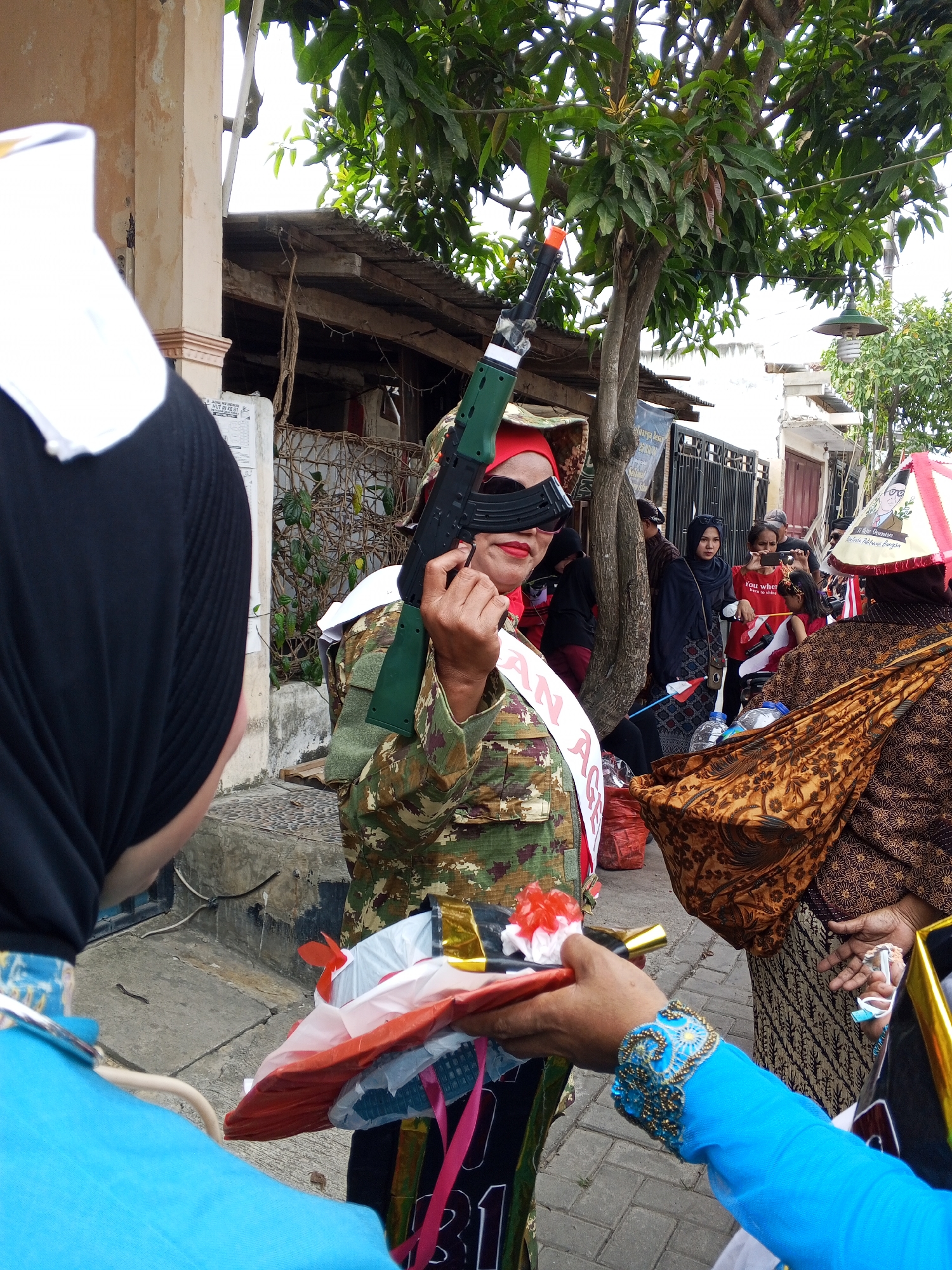 In the city of Ciruas, Indonesia, a person dressed in a camouflage outfit and a red headscarf is holding a green toy rifle. They are also wearing dark sunglasses and a sash that reads "AN AGE". The individual is part of a procession or event, as other people are visible in the background, some of whom are dressed in traditional attire. One person in the foreground is wearing a bright blue outfit and appears to be holding out a gift or offering. The environment suggests an outdoor street scene, with buildings and trees visible. The overall atmosphere seems festive or ceremonial, given the costumes and the gathering of people.