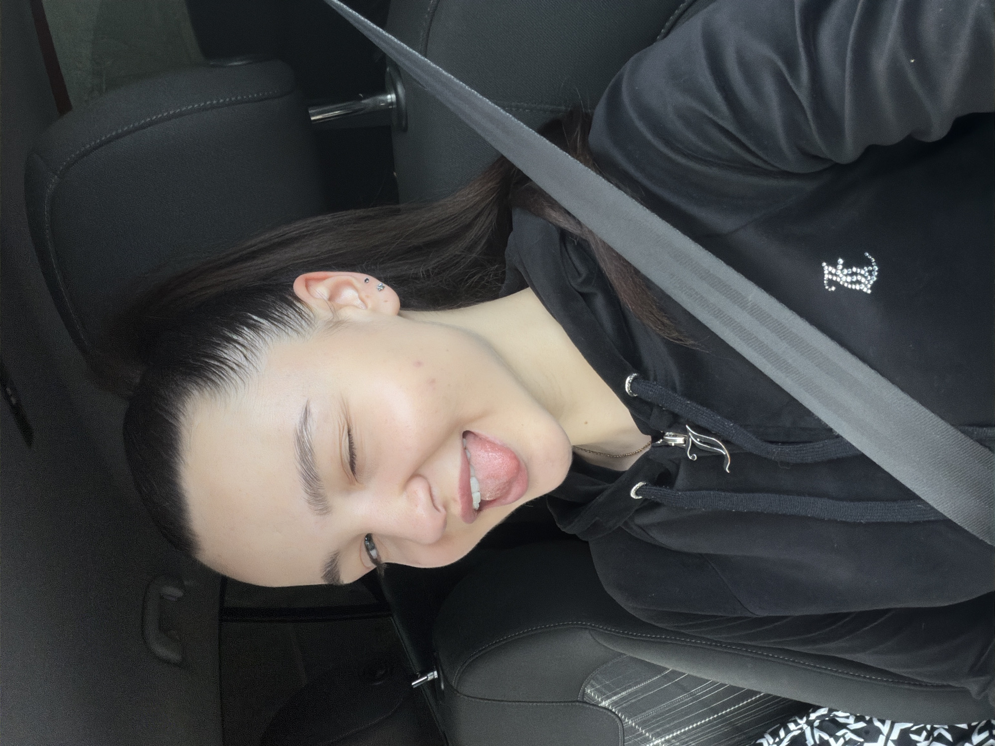 A young woman with dark hair is in the driver's seat of a car. She is wearing a black hoodie with a silver-colored design on the front and has a seatbelt across her torso. She is sticking her tongue out and partially closing one eye. The car's interior is dark gray leather. The location is Busmantsi, Bulgaria.