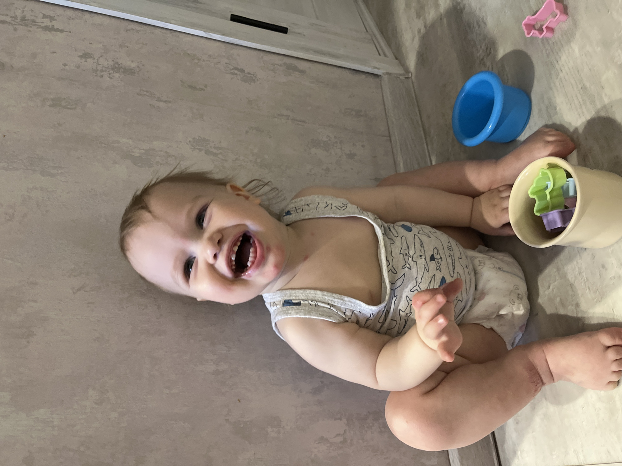 An indoor scene in Chaikovsky, Russia, depicts an infant seated on a light-colored floor against a textured, pale gray wall. The infant, with light brown hair and fair skin, is smiling broadly with an open mouth, revealing several teeth. Red marks are visible on the chin and around the mouth. The infant wears a light gray sleeveless bodysuit adorned with blue shark and fish patterns, and a white disposable diaper. The left arm is bent, and the left hand rests on the floor. The right arm is also bent, with the hand partially visible. The legs are bent, with feet flat on the floor. To the infant's lower left, a beige plastic cup holds multiple colorful plastic cookie cutters, including green and purple shapes. To the infant's lower right, a small, inverted blue plastic cup is visible, alongside a pink plastic animal-shaped cookie cutter lying on the floor. A white door frame with a vertical black handle is present in the background on the right.