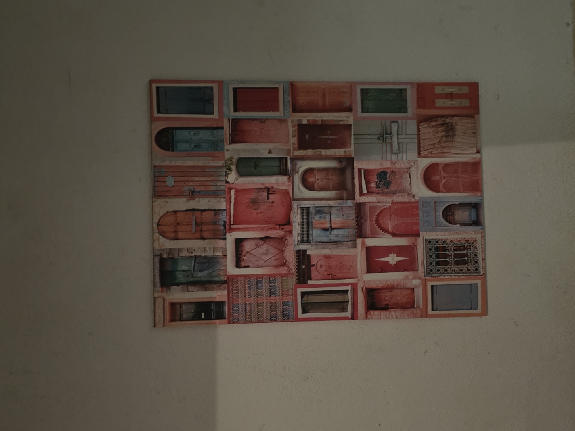 A rectangular artwork, composed of a grid of multiple images of doors, hangs on a light-colored, possibly off-white or beige, wall. The artwork is positioned slightly to the right of the center of the frame, leaving a significant plain wall space to its left.
The collage features a variety of doors, each depicted within its own rectangular or square segment. The doors display diverse architectural styles, colors, and textures, suggesting different origins and ages. Colors include shades of red, pink, blue, green, orange, and brown. Some doors appear weathered and rustic with peeling paint and distressed surfaces, while others show more intricate designs, such as arched doorways, patterned grilles, or decorative paneling. Examples include a bright red door, a teal-colored door, a door with a pointed arch, and several with decorative metalwork or wooden patterns.
The room is indoors, indicated by the plain wall and the presence of the artwork. The lighting is somewhat dim, casting subtle shadows on the wall, particularly below and to the right of the framed piece. No specific activity is taking place; the image is a static shot of the artwork. There are no visible people or discernible text in the image.