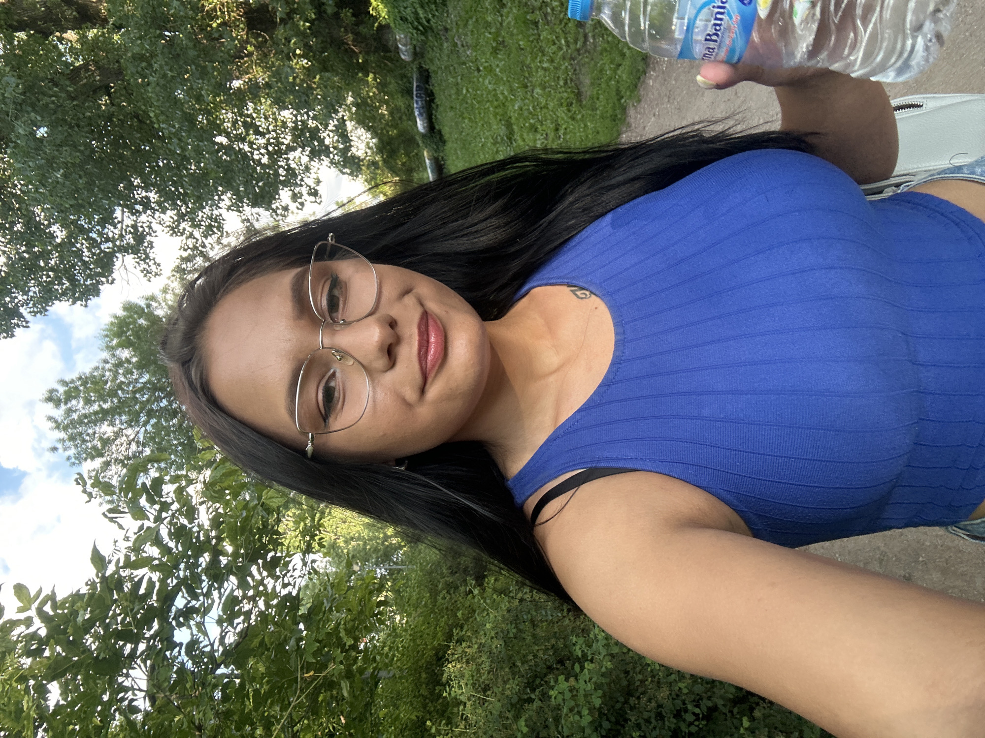 A young woman with long dark hair and glasses is taking a selfie outdoors. She is wearing a blue ribbed tank top. She is holding a bottle of water in her right hand. The background shows trees and greenery, suggesting a park or natural area within the city of Sofia, Bulgaria. The lighting indicates it is daytime.