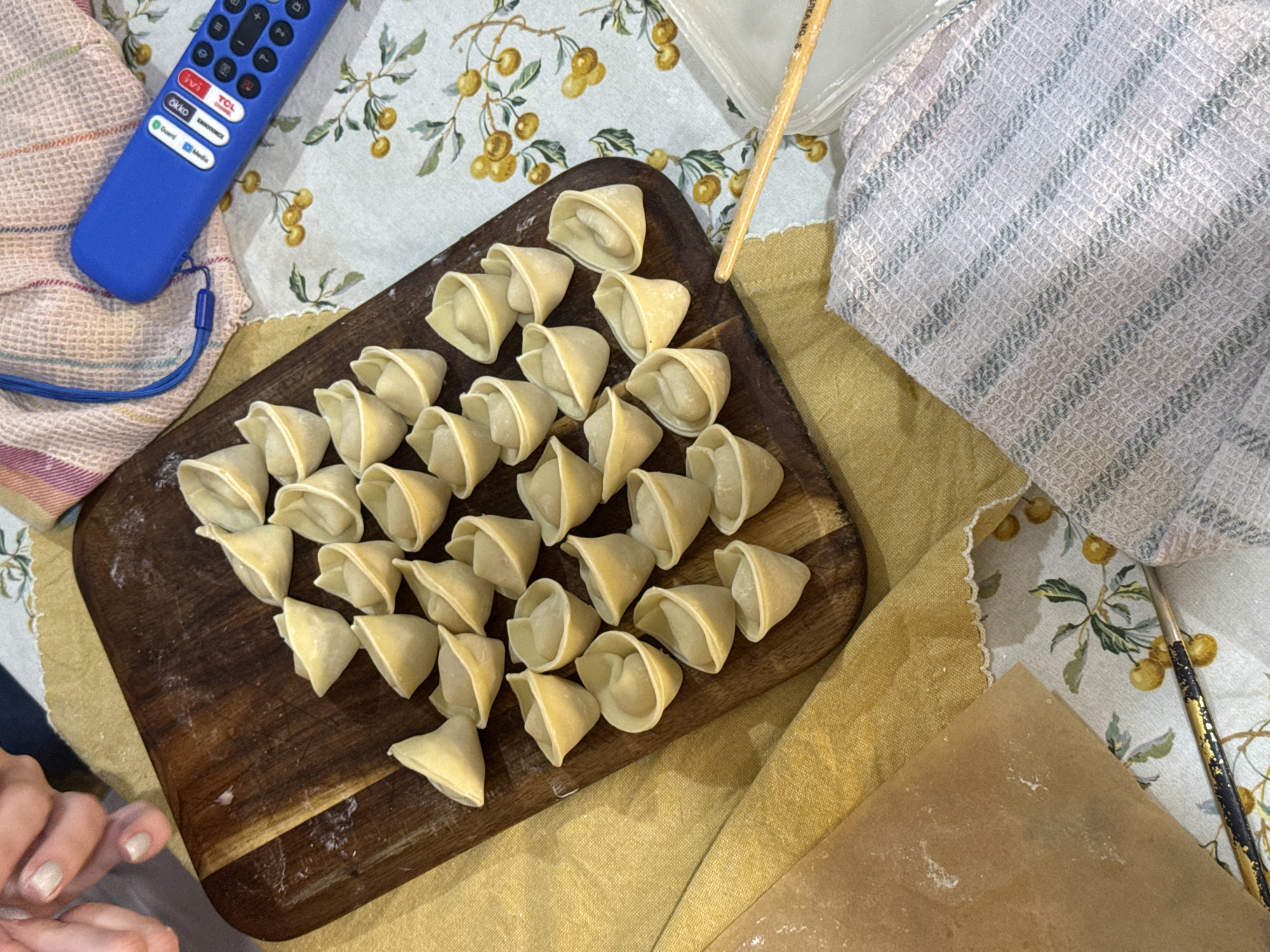 A high-angle shot shows a wooden cutting board filled with approximately 30 uncooked dumplings, likely pelmeni, arranged closely together. The board is set on a floral patterned tablecloth. To the left of the board lies a blue remote control next to a striped cloth. To the right is a checkered dish towel. A wooden stick and a piece of parchment paper with scattered flour reside on the lower right. A hand is visible in the bottom left corner. The scene suggests someone is preparing food, possibly in a home setting. The location is identified as Krasnoyarsk, Russia.