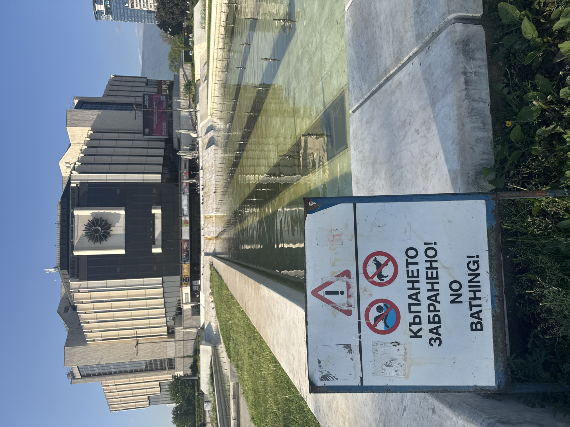 The image depicts a daytime scene in Sofia, Bulgaria, featuring the National Palace of Culture (NDK) in the background and a long, shallow reflecting pool in the foreground. A sign prohibiting bathing is visible near the pool's edge. The sky is clear and blue. A tall building is seen in the distance. Grass and greenery border the pool.