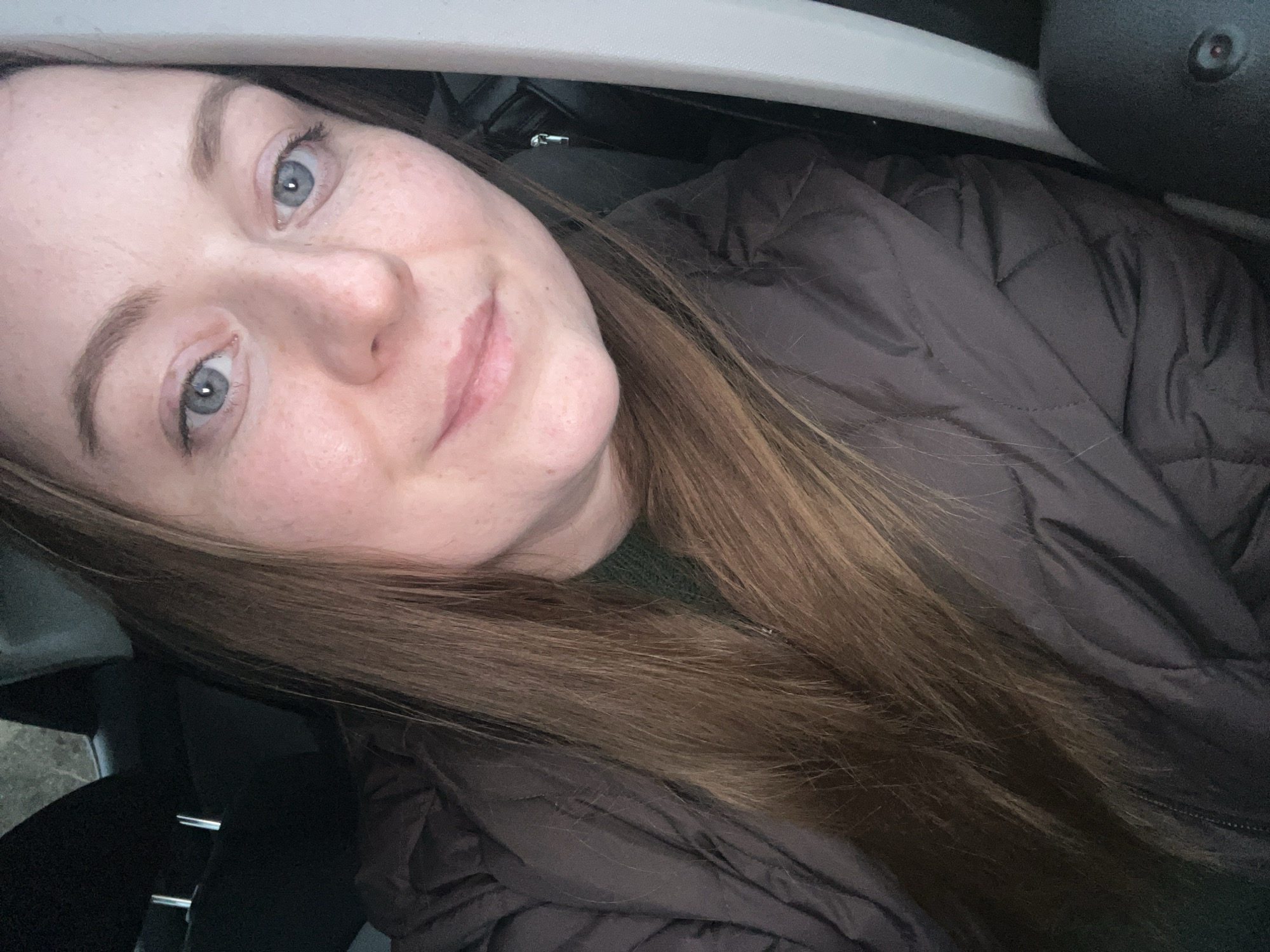 A medium-close-up shot depicts a woman with light skin, freckles, and blue eyes, looking directly at the camera with a slight smile. She has long, brown hair. She is wearing a brown quilted jacket and a dark green garment underneath. The setting appears to be inside a car, based on the visible car interior elements in the background, including a seatbelt and part of the car's ceiling. The location in the city of Раздолье, Russia cannot be ascertained from the image.