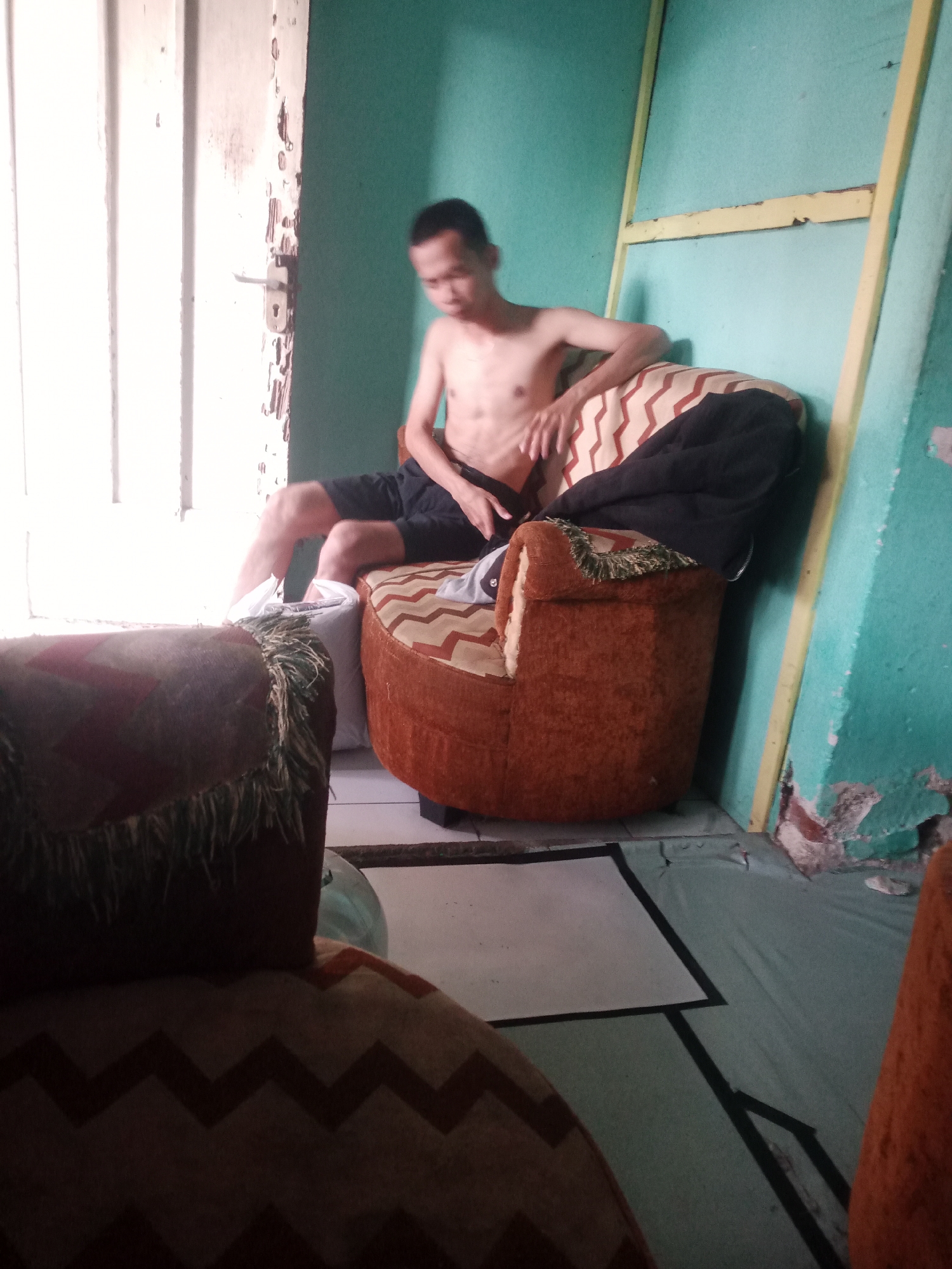 In Tasikmalaya, Indonesia, a man is seated on an armchair in a room. The man is shirtless and wearing dark shorts, with his legs crossed. The armchair is upholstered in a brown fabric with a zigzag pattern, and it is placed near a door that appears to be ajar. The room has a turquoise-colored wall and a wooden frame structure leaning against it. The floor is tiled, and a white mat with a black border is visible. In the foreground, another armchair with a similar zigzag pattern is partially visible. The lighting in the room is dim, with natural light entering from the doorway. The scene suggests a casual, indoor setting.