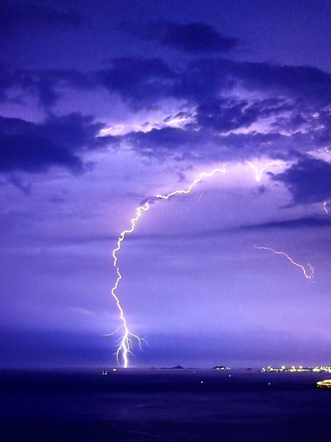 A dramatic lightning strike illuminates a dark, stormy sky over a body of water. The lightning bolt, a vibrant white with yellow accents, forks out into a complex pattern as it descends towards the water's surface. Dark purple clouds, heavy with rain, dominate the upper portion of the image, creating a stark contrast with the bright flash. The water below is a deep, muted blue, reflecting the dark sky. In the distance, along the horizon, a faint line of lights suggests a city or town. There are no visible people or animals in the image. The scene captures a powerful natural event occurring over a coastal landscape, possibly near Kafr Yasif, Israel, given the context.