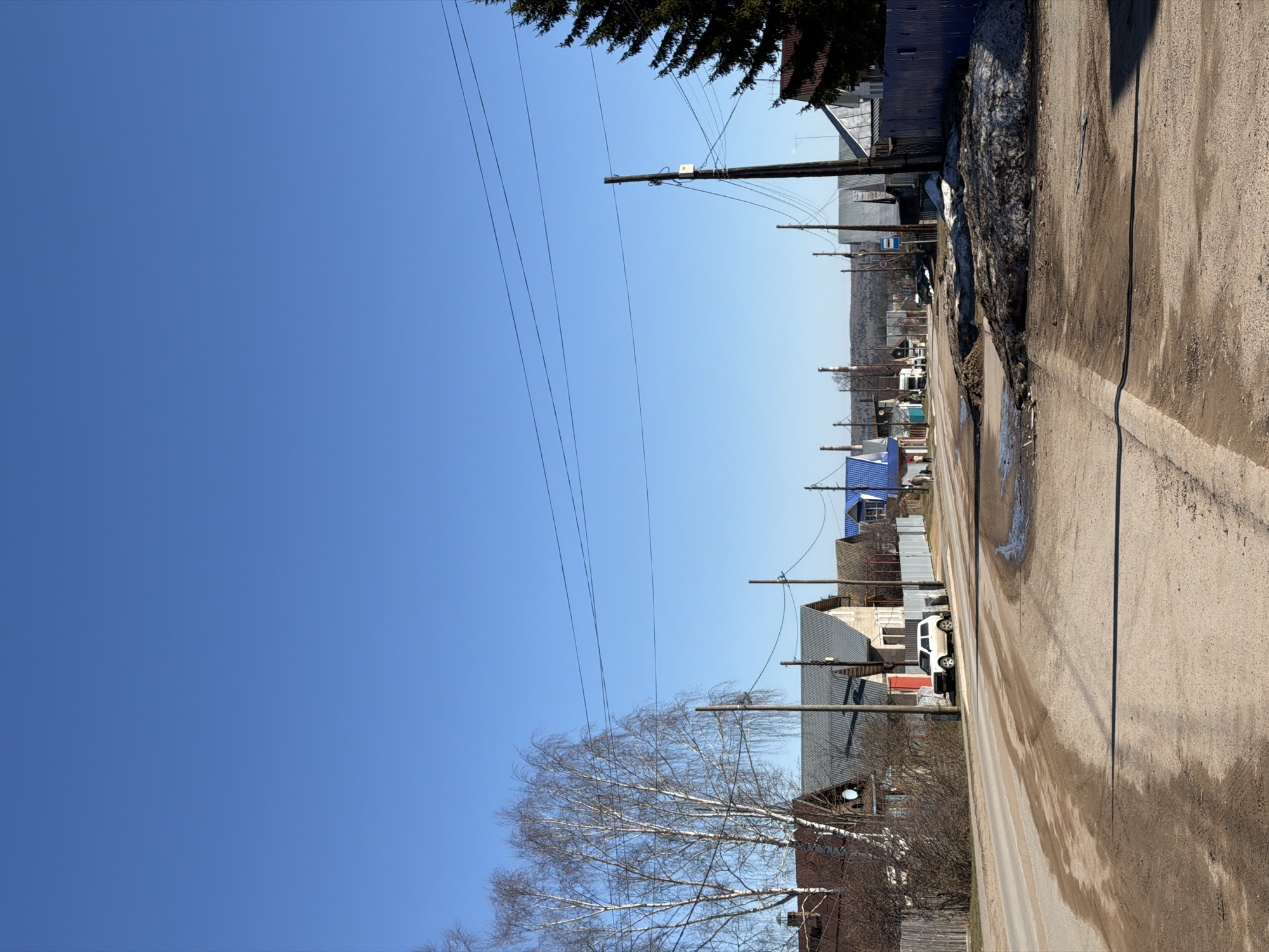 An eye-level, sunny view of a street in Chaikovsky, Russia. Power lines stretch along the length of the street, connecting wooden telephone poles. A two-lane asphalt road is visible, dirty and wet in places. Houses are on either side of the street, with varying colors and construction styles. Some roofs are gray, some blue. A white car is parked along the right side of the road. Patches of snow still remain on the side of the road. Trees are leafless near the bottom of the frame, while a pine tree is near the top right corner of the frame. The sky is a clear, bright blue.