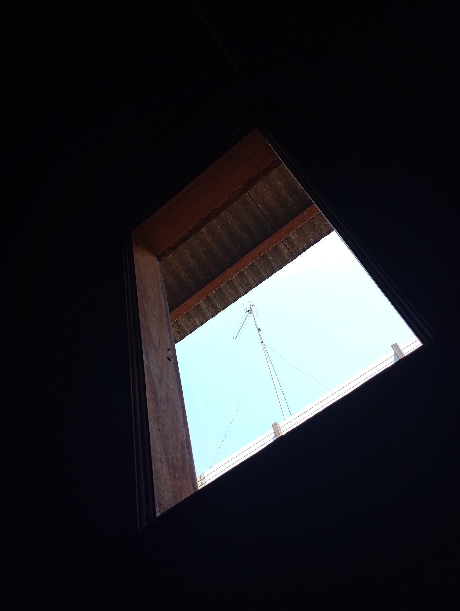 The image captures a view from within a dark interior, looking upwards and outwards through a rectangular opening in what appears to be a roof or ceiling. The opening is framed by wooden beams and corrugated roofing material. Beyond the opening, a bright, clear sky is visible. In the sky, a tall, multi-element antenna is prominently featured, mounted on a pole and supported by guy wires. The antenna is positioned slightly to the left of the center of the frame. The overall impression is of looking out from a sheltered space into the open air, with the antenna being the primary man-made object in the outdoor scene. The location is identified as Kecamatan Haurgeulis, Indonesia, suggesting this is likely a residential or utility structure within that area.