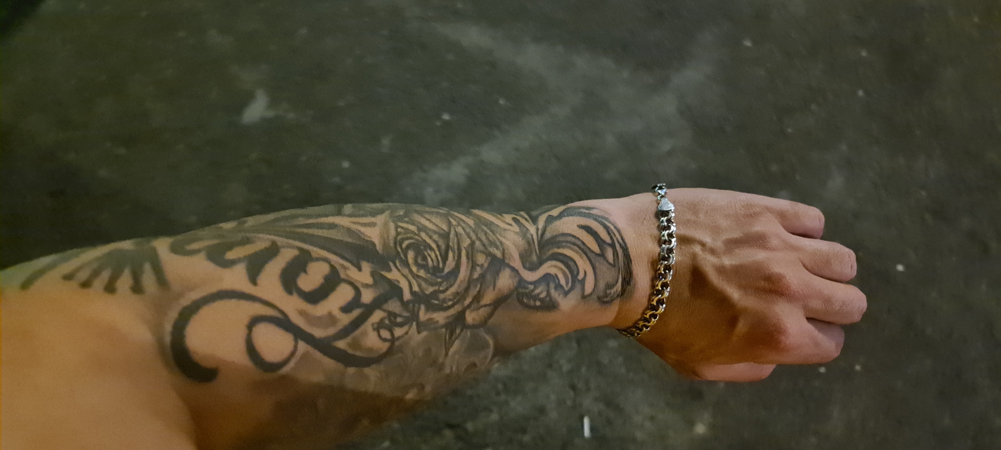 The media file shows a close-up view of a person's tattooed arm and hand, adorned with a silver bracelet. The arm features intricate black ink designs, including a prominent rose and swirling patterns. The bracelet is made of interlocking links and is worn on the wrist. The background is a dark, textured surface, possibly concrete or asphalt, with some scattered lighter speckles.
The lighting suggests it might be daytime, but the overall tone is dim. There are no clear environmental cues to indicate the specific location within Shaki, Azerbaijan, beyond the context provided. The image is focused on the details of the tattoo and the bracelet, implying an interest in personal adornment and body art. No specific activity is depicted, as the focus is on a static pose. The image does not contain any visible text.