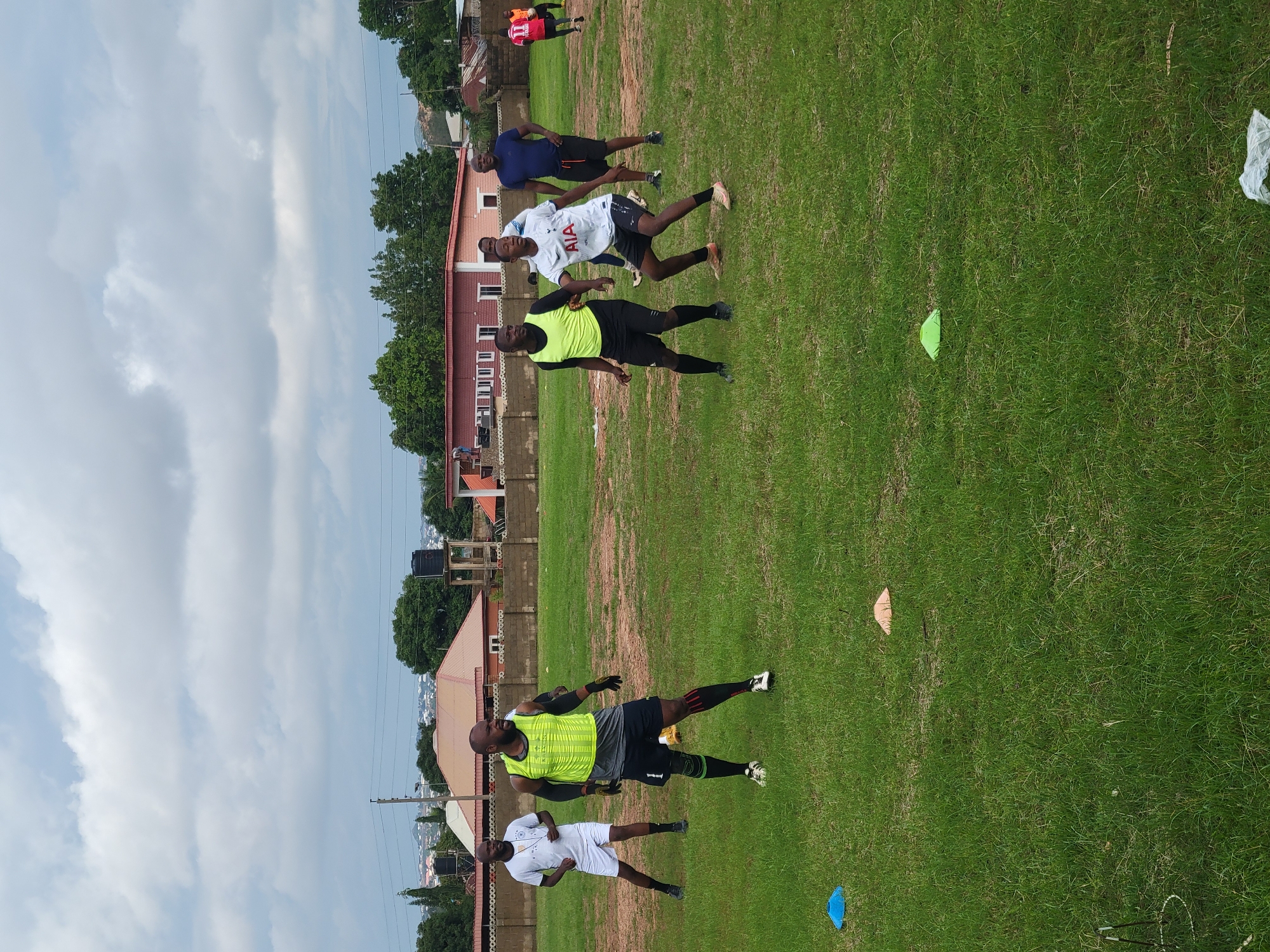 The image depicts a group of men engaged in a football training session on a grassy field. The setting appears to be an open area within a city, likely Jos, Nigeria, given the architectural style of the buildings in the background, which include houses with red tiled roofs and some featuring water tanks. The sky is overcast with thick, grey clouds.
In the foreground, several individuals are visible in athletic attire. One man in a yellow vest and black shorts is in focus, positioned centrally, appearing to be in a defensive stance. To his left, another man in a white t-shirt and white shorts is also in a ready pose. Further to the left, a man in a white t-shirt and white shorts stands with his hands on his hips, observing. In the middle ground, another man in a white t-shirt with "AIA" printed on it and black shorts is in motion, seemingly interacting with the man in the yellow vest. To the right of this group, a man in a blue t-shirt and black shorts stands looking towards the training activity. In the background, a man in a red jersey with the number "11" is also present on the field.
Scattered across the field are several colored cones, likely used for drills, including blue, orange, and green ones. The grass appears to be a typical field turf, with some patches of dry earth visible. The overall atmosphere suggests a casual yet focused training environment, possibly at a local sports ground. The presence of buildings and infrastructure in the background indicates an urban or semi-urban location.