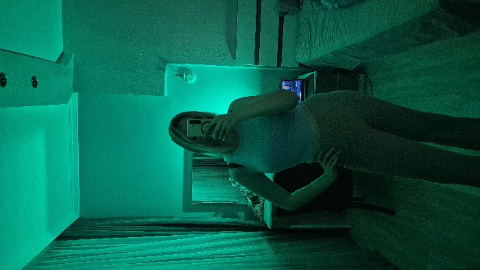 A woman takes a selfie in a green-lit room. She holds a phone up to her face, obscuring it. She wears a light-colored t-shirt and leggings. A bed and some furniture are visible in the background. The room appears to be illuminated with green neon lights, which cast a monochromatic hue over the entire scene.