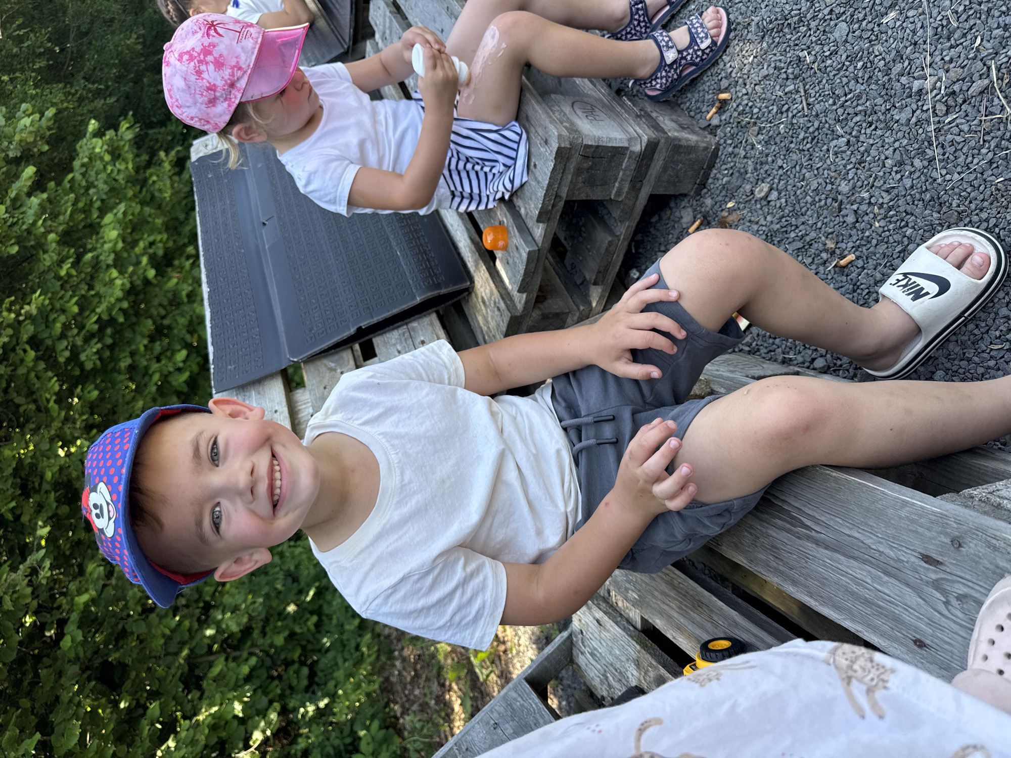 A medium shot captures two children sitting on a wooden bench outdoors. In the foreground, a young boy with blond hair wears a blue baseball cap with a red brim, a white t-shirt, and gray shorts. He is smiling and looking at the camera. To his right, a young girl with blonde hair tied in a ponytail wears a pink cap with a palm tree design, a white t-shirt, and a striped skirt. She is applying sunscreen to her leg. A yellow toy truck is partially visible on the bench to the boy's left. The background features green foliage and a dark, textured object. The ground is covered in grey gravel with several cigarette butts scattered around. The scene appears to be in a park or outdoor recreational area. The location is Beltershain, Germany.