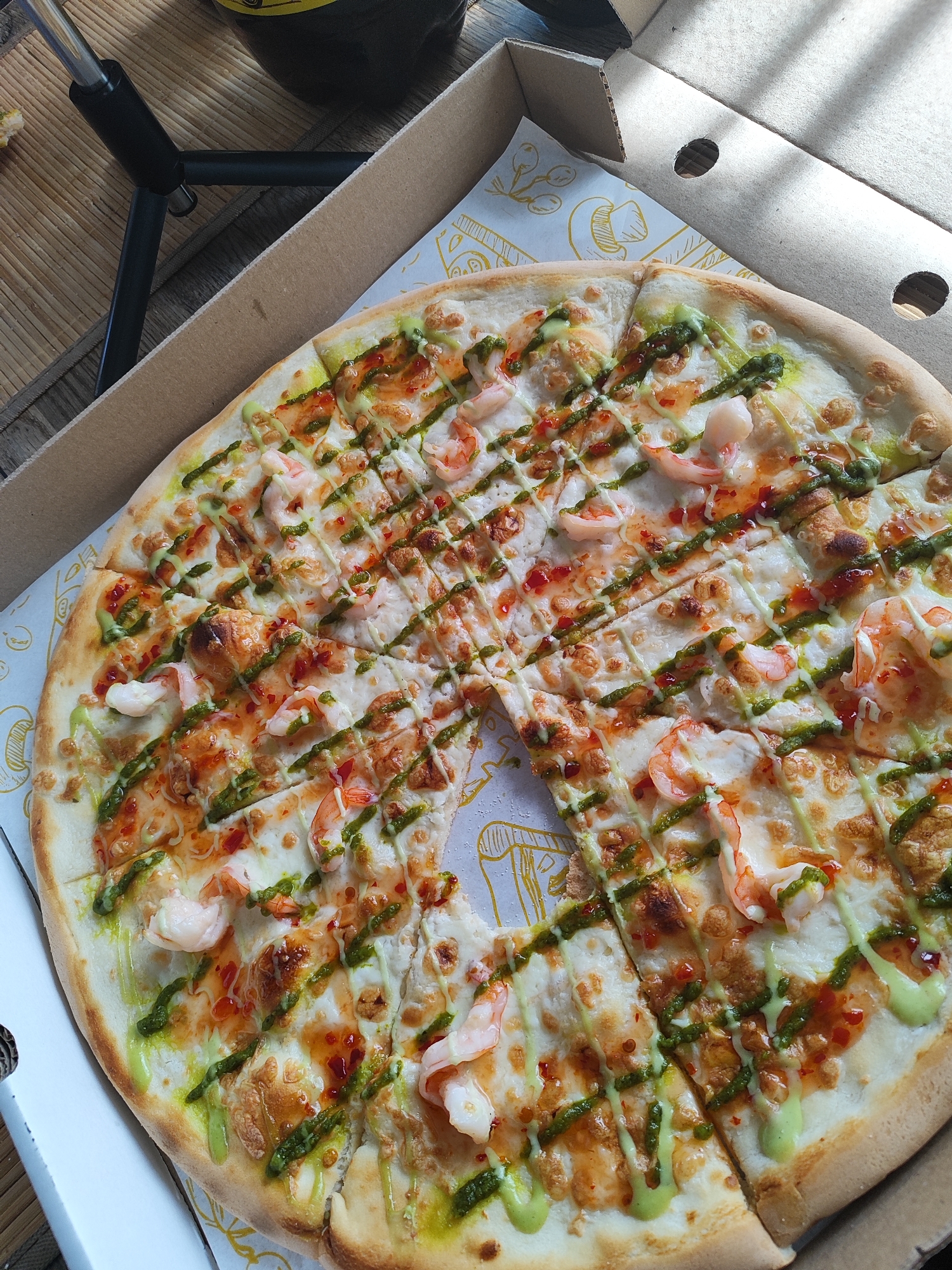 A high-angle, overhead shot depicts an opened pizza box containing a shrimp and sauce-drizzled pizza, situated on a textured surface. The circular pizza, missing one slice from its right side, is sliced into multiple rectangular portions. Its toppings include whole cooked pink shrimp, a generous amount of melted white cheese, a crisscross drizzle of vibrant green sauce (possibly pesto), and an intersecting drizzle of reddish-orange sauce (possibly chili oil or a similar condiment), with scattered small brown granular elements.
The pizza rests on a white paper lining inside a light brown cardboard pizza box. This paper lining features yellow line-art illustrations, including mushroom motifs. The box lid is partially open, revealing its underside and two circular cut-outs.
To the upper left of the pizza box, a black, collapsible tripod is visible on a woven light brown mat, which itself rests on a darker, planked wooden surface. A dark-colored bottle with a yellow label element is partially visible in the upper right quadrant of the image. The scene is illuminated by light from an unseen source, casting distinct shadows from the pizza box lid and the tripod. No individuals are present in the image. The specific location in the city of Marusino, Russia, cannot be determined from the visual information provided.