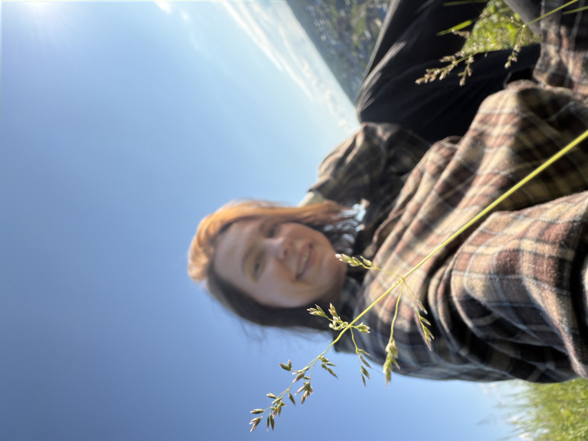 The image displays a low-angle shot of a young person outdoors against a bright blue sky. The main subject, appearing to be a female with reddish-brown hair, is seen from a slightly elevated perspective, looking up at the camera with a gentle, relaxed smile. She is wearing a plaid shirt, featuring patterns in shades of brown, beige, and possibly dark blue.
A stalk of grass with tiny seed heads is prominently featured in the foreground, adding a natural element to the composition. The background is dominated by a clear, vibrant blue sky, indicating daytime and sunny weather. In the upper left portion of the image, blurred hints of a distant landscape with green foliage or structures can be observed, suggesting an open, possibly rural or elevated outdoor location. The overall mood is serene and positive, with the subject appearing content in the natural environment. The image has a soft focus, giving a slightly dreamy quality to both the foreground elements and the person. There is no visible text. The context places the location in Ольховка, Russia.