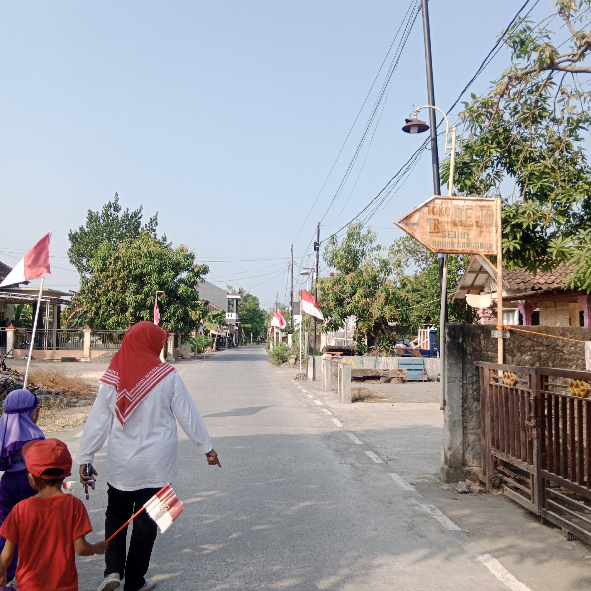 This image depicts a street scene in Tegowanu Kulon, Indonesia, likely during a patriotic celebration given the presence of numerous Indonesian flags. In the foreground, a woman wearing a red hijab and a white shirt with red trim walks away from the camera, holding a small Indonesian flag and pointing to the right. Beside her, a young boy in a red shirt and red cap walks with another small Indonesian flag. Another child, partially visible, is dressed in a purple hijab. The street is paved and has a dashed white line marking the center. On the left side of the street, houses with Indonesian flags displayed are visible, along with trees. On the right side, a rusty metal gate stands next to a wall. A directional sign pointing left reads "TOKO REJO" and "SEDIA BAHAN BANGUNAN," indicating a shop selling building materials. Overhead, power lines crisscross the clear blue sky, and a street lamp is mounted on a pole near the sign. The overall environment suggests a residential area on a sunny day.