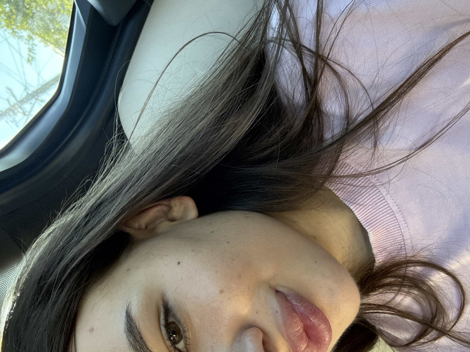 The image shows a close-up of a young woman's face and upper torso, taken from a side-view perspective. She has long, dark brown hair that falls across her face and shoulders. Her skin appears to have a few freckles and a slight rosy hue on her cheeks. She is wearing a light pink, long-sleeved shirt with a ribbed neckline. Sunlight is hitting her face, casting a soft glow. Her left eye is visible, with brown irises. Her lips are full and slightly parted, appearing glossy. The background shows a car's interior, with a black trim on the top and a light-colored seat. Through a window, a bright sky with some power lines and greenery can be seen, suggesting the car is in motion or parked outdoors. The photograph was taken in Zorleni, Romania.
