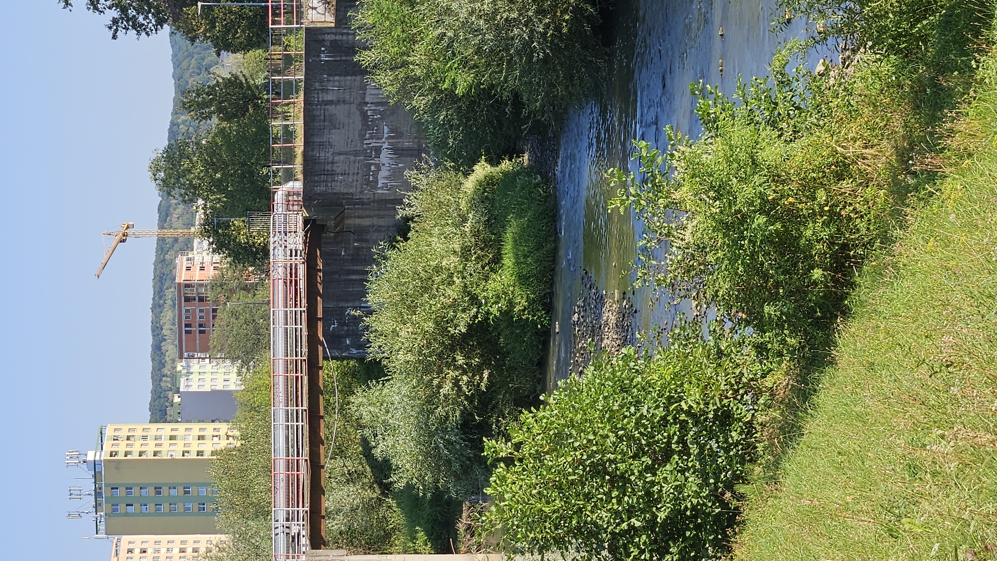 The image depicts a river flowing through a city, with a bridge spanning across it. In the background, several apartment buildings and a construction crane are visible, suggesting an urban environment. The riverbanks are lined with lush green vegetation, including trees and bushes. The water appears calm, with a rocky bed visible in some areas. The sky is clear and blue, indicating a sunny day. The overall scene suggests a blend of urban development and natural scenery. The location is identified as Prešov, Slovakia.