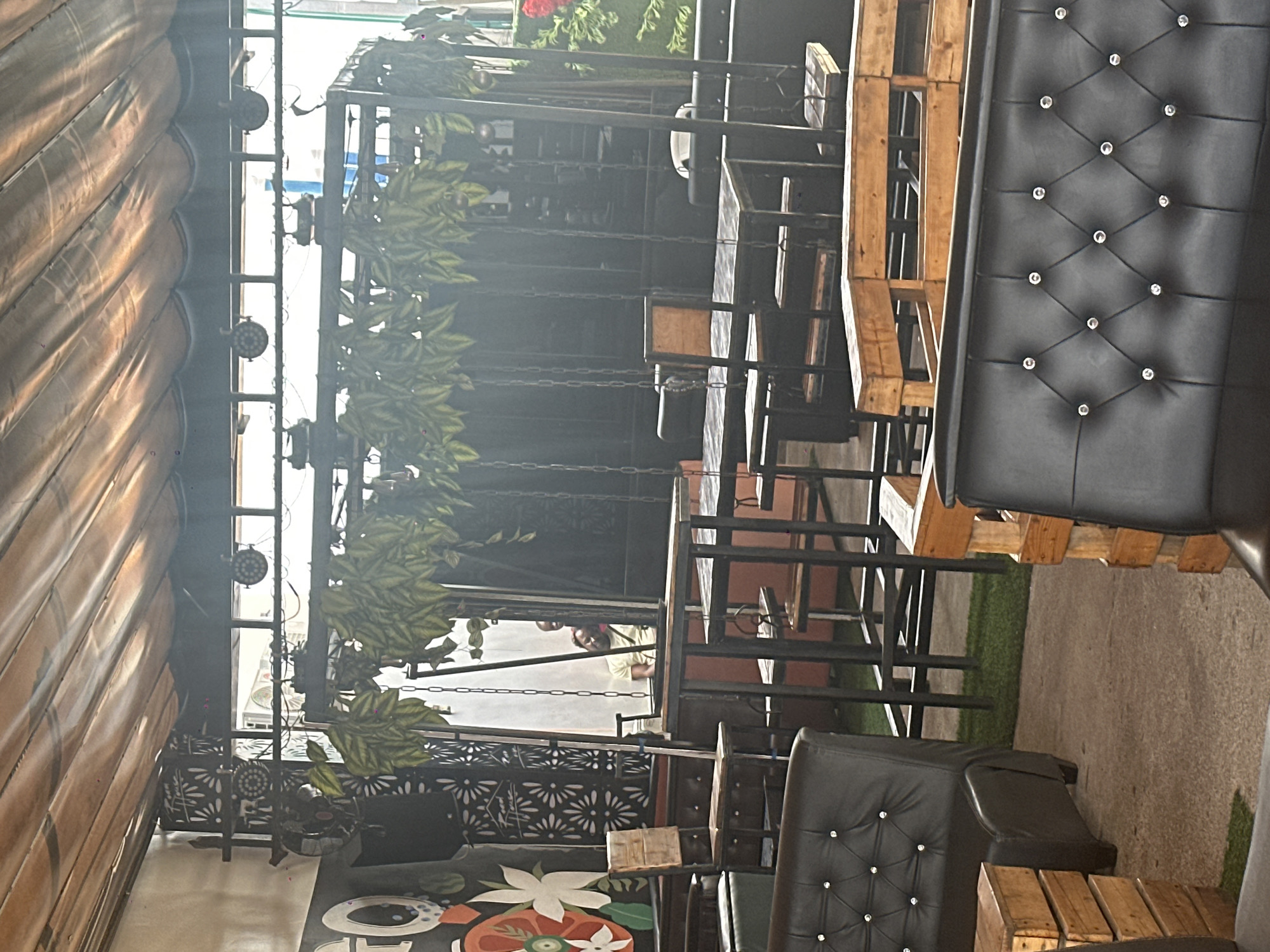 The image depicts an outdoor seating area, likely a cafe or restaurant, in Howayi, Nigeria. The setting appears to be a covered patio, with a striped awning extending from the left side of the frame. Several tables and chairs are arranged, with some made from repurposed wooden pallets. Black leather sofas with diamond tufting are also visible, adding a touch of comfort and style to the seating.
The scene is illuminated by artificial lights, suggesting it could be evening or an indoor space with artificial lighting. The background shows a dark structure with hanging chains and artificial greenery, possibly decorative elements or part of a stage setup. In the middle ground, a person is partially visible, wearing a light-colored top. The overall ambiance seems relaxed and casual, suitable for dining or socializing. The ground is a mix of concrete and what appears to be artificial turf. There are no discernible emotions conveyed by the people present. No visible text is present in the image.