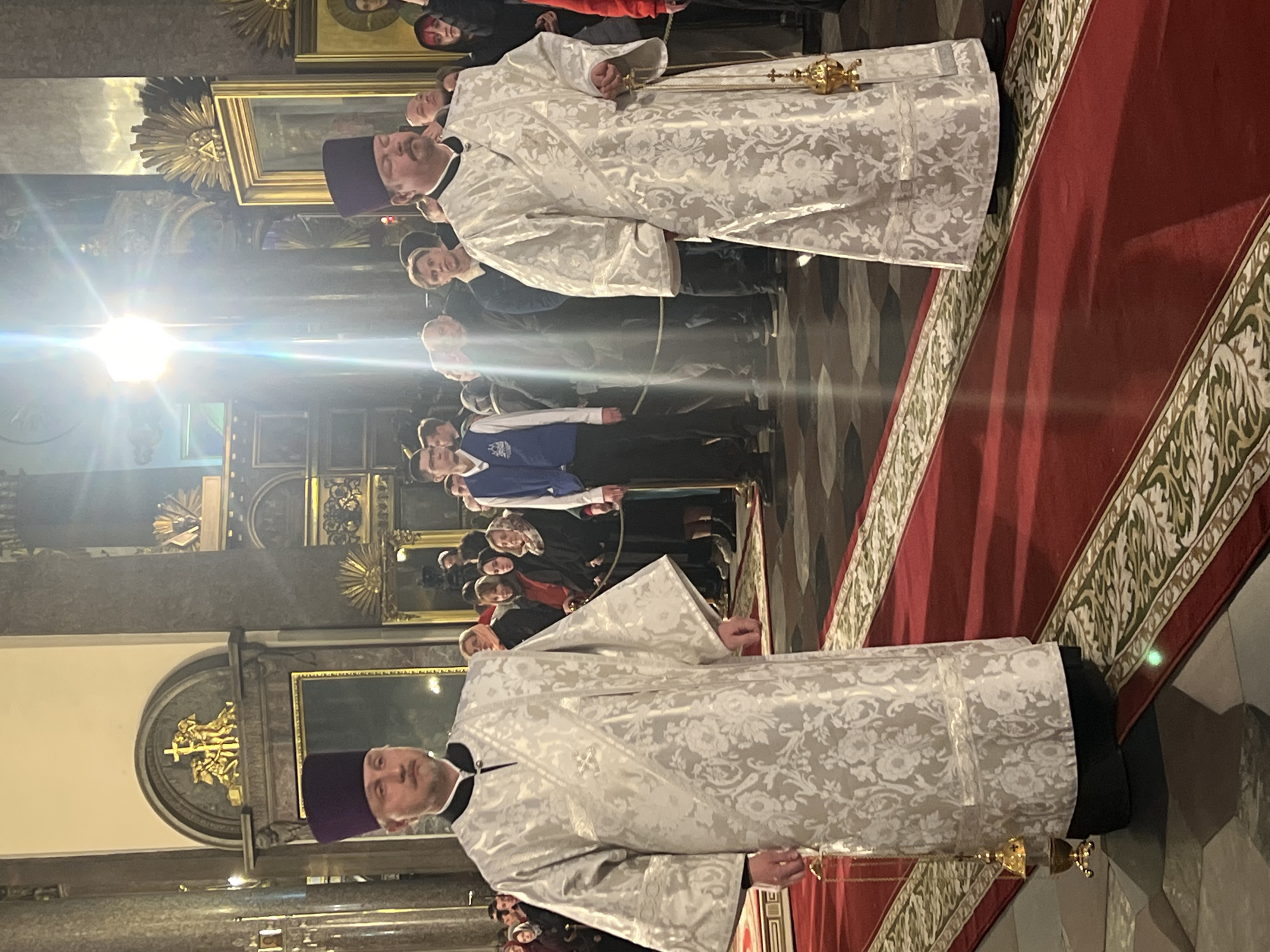 The image depicts two Orthodox priests in a church. They are wearing white, patterned vestments and purple hats. Each is holding a golden ornate object with small bells. A crowd of people stands behind them, some wearing blue and white attire. The setting is ornate, featuring gold-framed religious paintings on the wall. A red carpet with patterned edges extends along the floor. The church is identified as located in Saint Petersburg, Russia.
