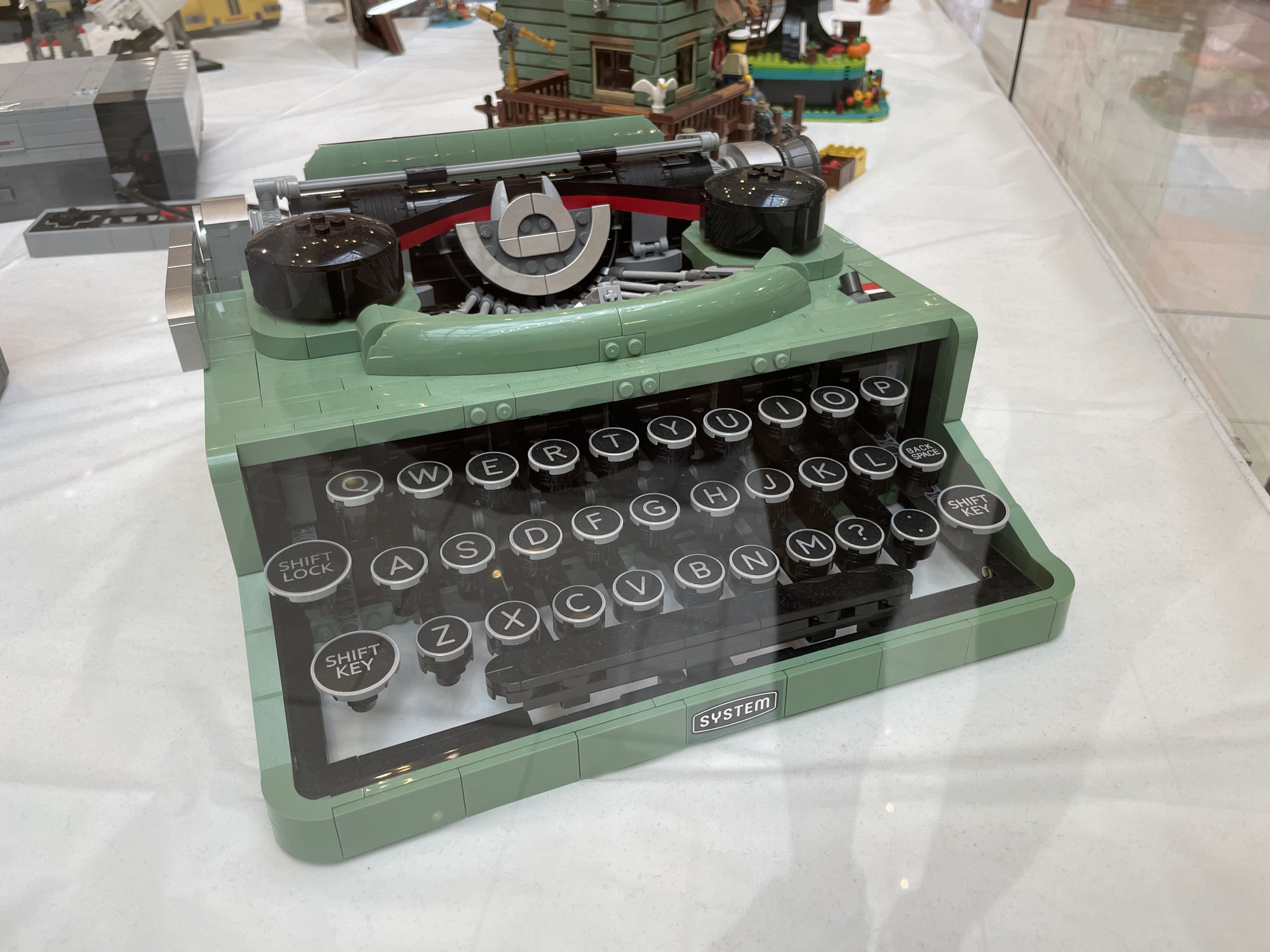 The image shows a detailed Lego model of a vintage green typewriter. The typewriter is the main subject, with its keys, ribbon mechanism, and paper roller visible. The keys are circular with white lettering. The background is blurred and appears to be a white surface, likely a display table at an event. There are other Lego creations in the background, including a grey console and a brick building with figures. The scene is indoors, and the lighting suggests a well-lit exhibition space. The location is not specified in the image, and there is no indication of Sofia, Bulgaria.