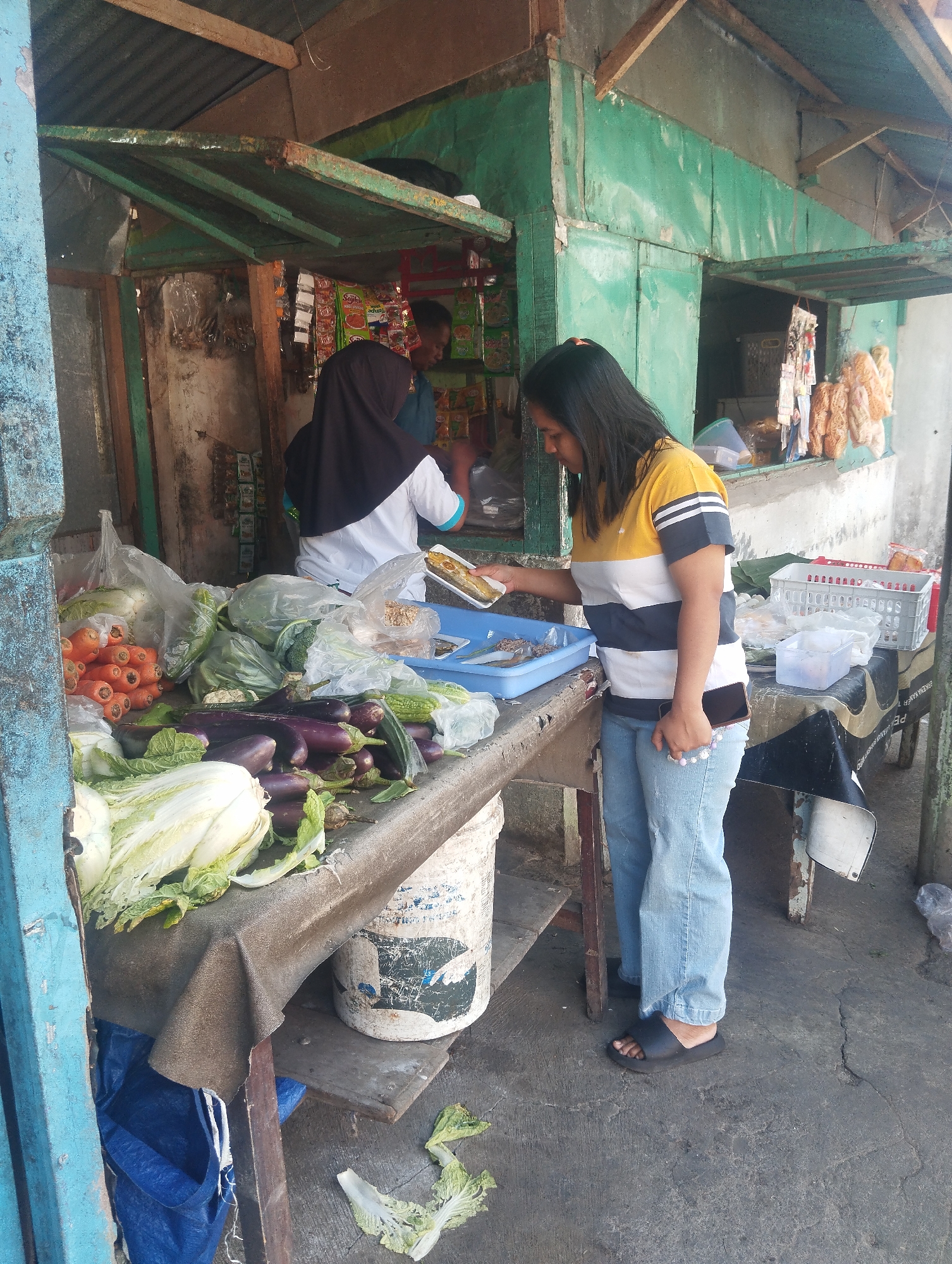 The image depicts a roadside stall in Malang, Indonesia, likely a fruit and vegetable market. In the foreground, a woman wearing a striped yellow, white, and black t-shirt and light blue jeans stands by a table laden with fresh produce. She is holding a mobile phone in her right hand and reaching with her left hand towards a blue tray on the table, which contains what appears to be fried food items.
The table is covered with a brown tarp and displays a variety of vegetables, including carrots, eggplants, napa cabbage, bitter melon, and leafy greens. Some of the produce is in plastic bags. Behind the woman, another person wearing a black hijab and a white shirt with blue trim is partially visible, seemingly interacting with the stall's wares.
The stall itself is a rustic structure with a corrugated metal roof and green painted wooden walls. Shelves inside the stall are stocked with packaged goods, likely snacks or small food items. The ground surface is concrete, showing some cracks and debris. The overall impression is of a busy, informal marketplace.