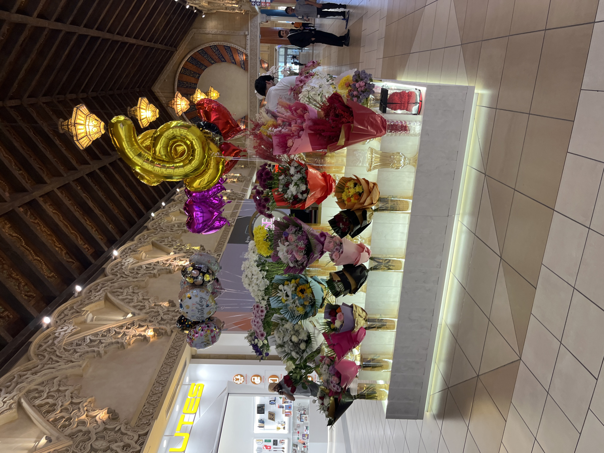 This is a medium shot, eye-level, full-color photograph taken indoors in what appears to be a mall or large retail establishment in Dubai, United Arab Emirates. The scene features a brightly lit flower display situated in the foreground, showcasing an assortment of bouquets arranged in vases of various sizes and materials, including glass and gold-toned cylinders. The flowers themselves are diverse in type and color, with prominent arrangements of red, pink, white, yellow, and purple blooms. Above the flower display, a collection of balloons is suspended, including heart-shaped balloons in red and purple, a cluster of colorful patterned balloons, and a large gold "6" shaped balloon.
In the mid-ground, several people are visible. On the left, a man wearing a turban and a light-colored shirt is standing near a retail store entrance, holding a red object, possibly a bouquet or a gift, and talking on a mobile phone. Behind the flower display, a person in a white t-shirt is attending to the flowers, their back to the camera. Further in the background, two men in business attire, one in a dark suit and the other in a lighter shirt and dark pants, are standing and conversing. Other shoppers and staff members are visible in the distance.
The environment includes a tiled floor with a geometric pattern in varying shades of beige and cream. The ceiling features intricate architectural details with ornate carvings and wooden beams, illuminated by decorative pendant lights with a warm glow. A store with a bright yellow "LUTES" sign is visible on the left side of the frame. The overall ambiance suggests a well-maintained and upscale retail setting.