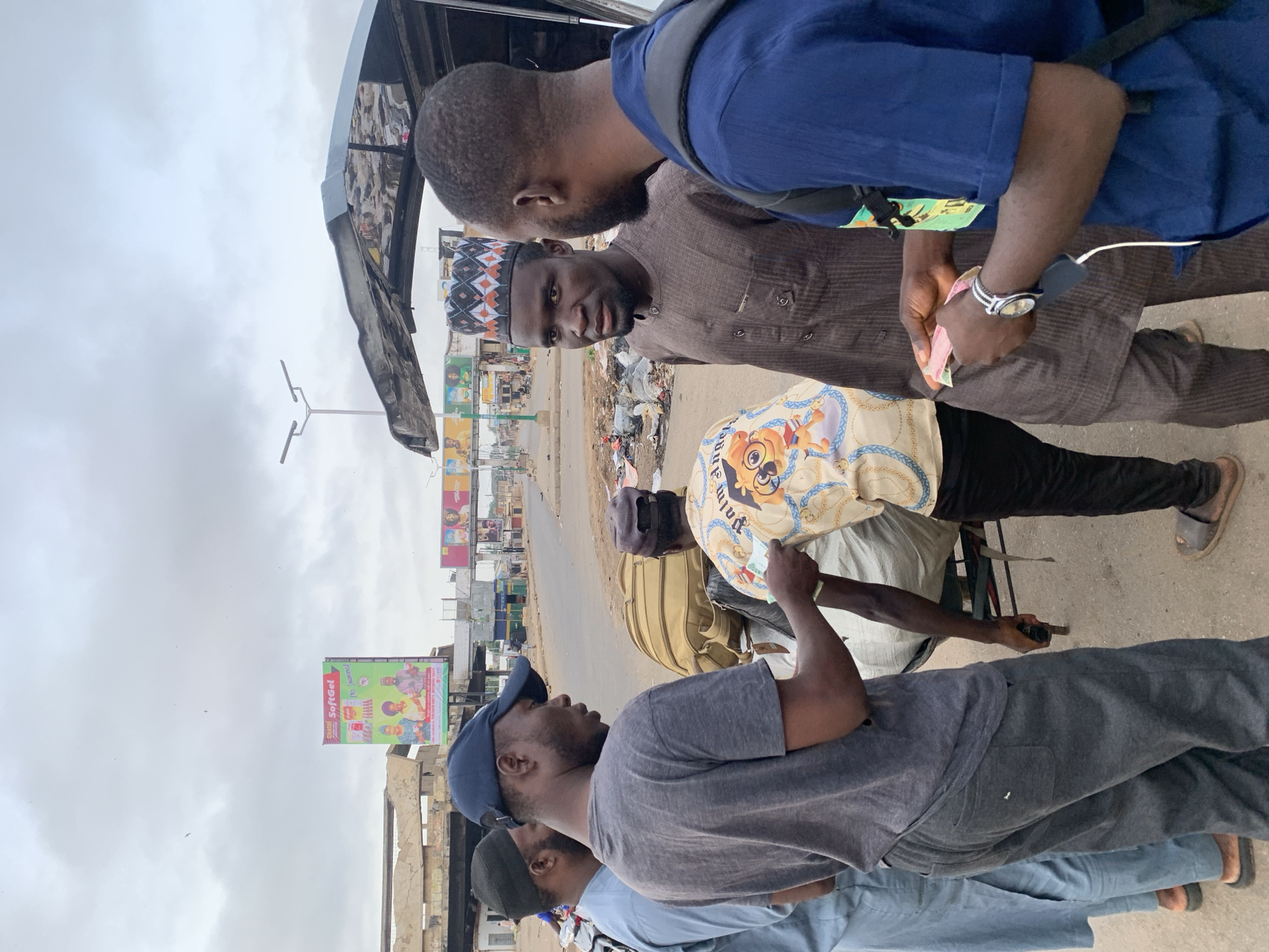 The image shows a street scene in Jos, Nigeria, with several men gathered around a vendor. One man is counting money. There are posters and billboards in the background, along with buildings. The environment appears to be urban, with evidence of litter and street infrastructure. The sky is overcast.