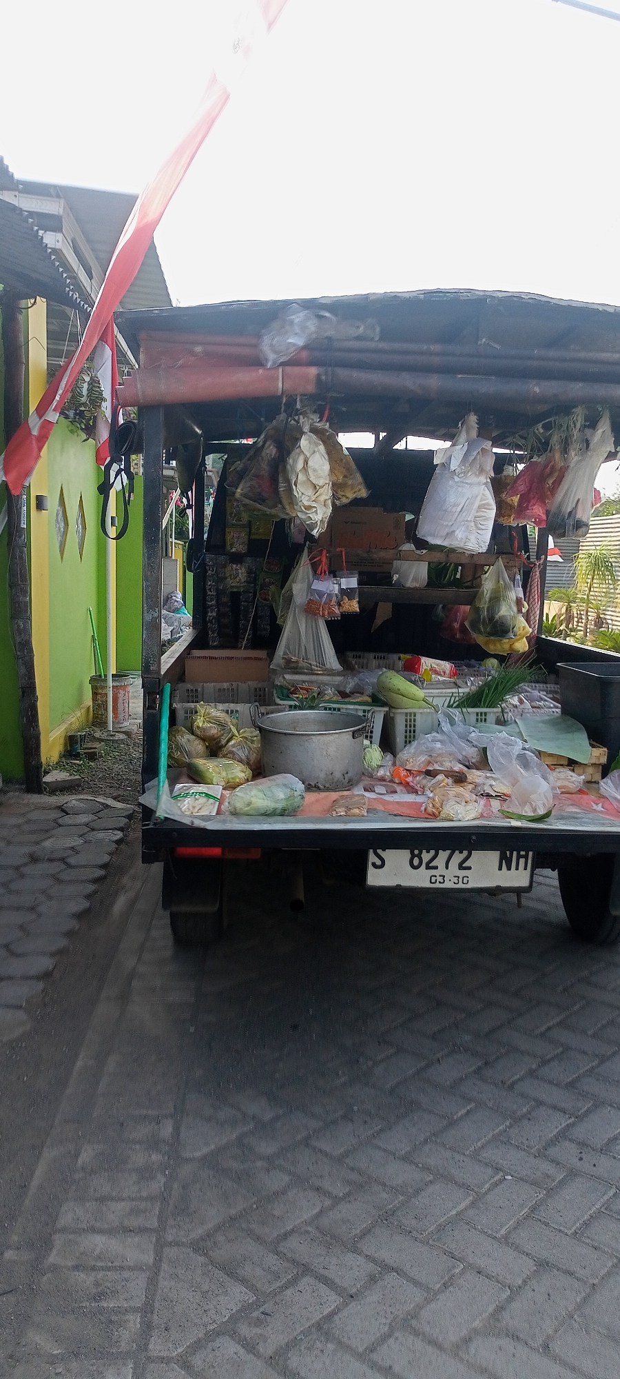 A low-angle, full-shot, outdoor, vertical image captured on a paved surface in Gempolklutuk, Indonesia, shows a mobile food stall on the back of a black utility vehicle. The stall displays a variety of food items and merchandise, including vegetables, packaged goods, and cooking utensils, all arranged on the vehicle's platform. The vehicle's license plate, "S 8272 NH," is visible at the rear. A red and white Indonesian flag is affixed to a pole on the left side of the stall. The scene is set against a backdrop of a yellow and green painted building with a metal roof structure supporting the stall. The ground is paved with interlocking concrete blocks, and shadows from the stall and surrounding structures are cast across the surface. The sky appears bright and overcast.