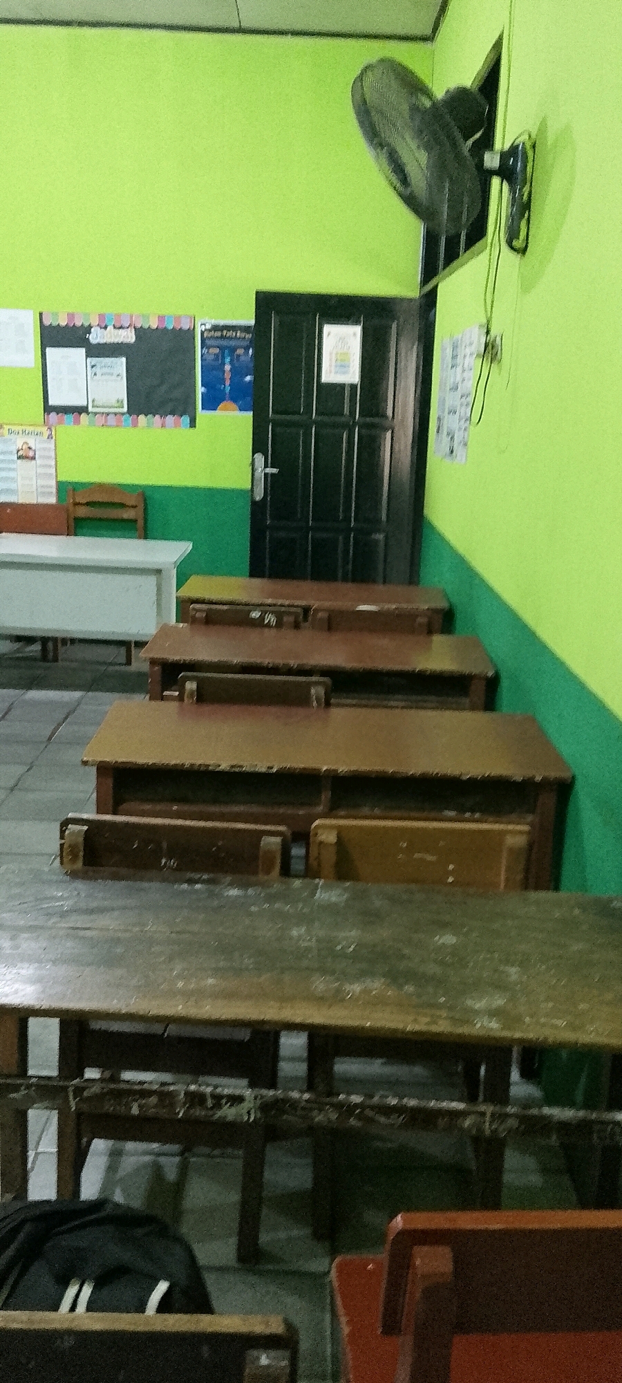 The image depicts a classroom scene, likely in Satriajaya, Indonesia, given the context. The walls are painted in alternating bands of lime green and dark green. A black oscillating fan is mounted high on the lime green wall. In the background, a dark brown door with a silver handle is visible, with a poster or notice attached to it. To the left of the door, a bulletin board displays various papers and notices.
The foreground and mid-ground are dominated by rows of wooden desks. The desks appear to be old and well-used, with some signs of wear and tear. The tops of the desks are a light brown color, while the lower structures are darker. Some desks have compartments for storage. A white cabinet or desk is situated to the left in the mid-ground, with a small wooden chair behind it. In the immediate foreground, the top of another long, dark wooden desk is visible, and part of a red chair is on the right. A black backpack with white straps is placed on the floor in the lower left of the image, resting on what appears to be a wooden bench. The floor is tiled with grey squares.