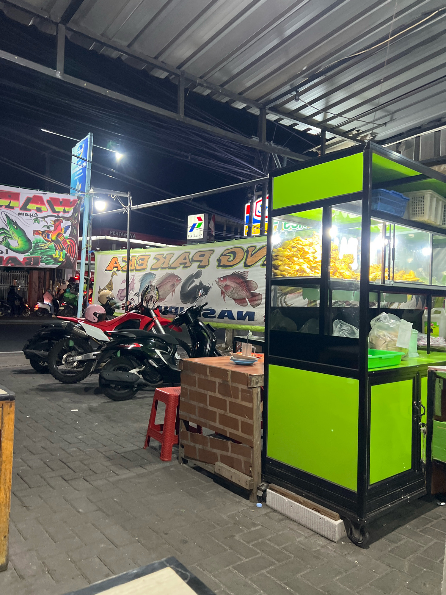 This image depicts a nighttime street scene in Denpasar, Indonesia, likely in an area with food vendors. The main focus is a brightly lit, lime green food cart with glass display cases filled with fried food. In front of the cart, there are two motorcycles parked, one red and one black, with helmets on them. To the left of the food cart is a small, brick-like structure with a red stool in front.
In the background, several large banners are visible. One banner on the left features illustrations of a shrimp and a rooster, with text that is partially obscured but appears to say "LAW". Another banner behind the motorcycles displays images of various fish and has large text that reads "PAK EKA". A sign for "PERTAMINA" gas station is also visible.
Several people are present in the background, suggesting a busy atmosphere, though their faces are not clearly visible. The scene is illuminated by streetlights and the lights from the food cart and nearby businesses, creating a lively, urban nighttime environment. The paving stones on the ground suggest a well-trodden public area. The overall impression is of a bustling local eatery operating at night.