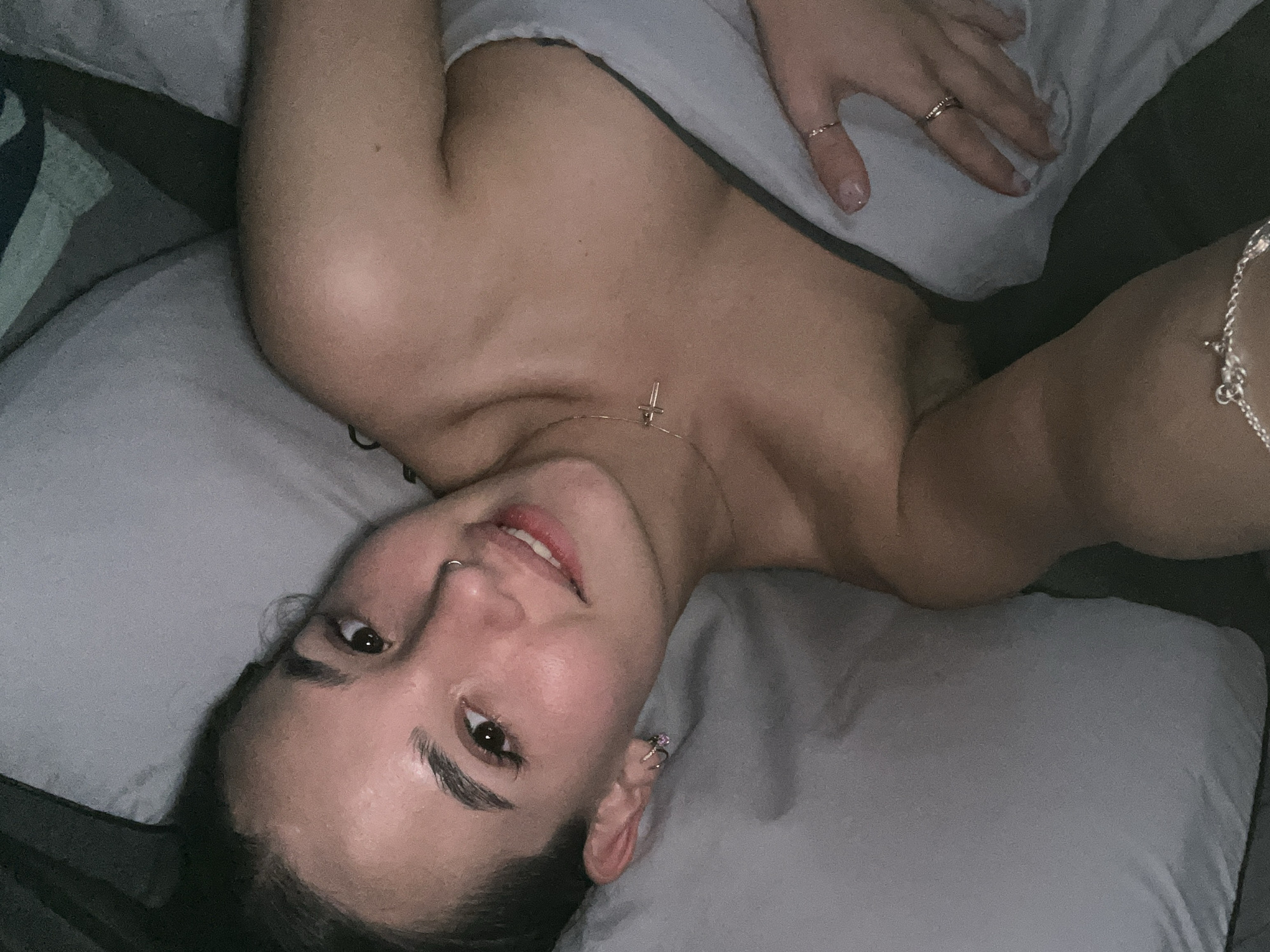 A person is lying in bed on a grey pillow and under a grey blanket. They have dark hair, light skin, and are wearing a nose ring, earrings, rings, and a necklace with a cross pendant. The location is not specified.