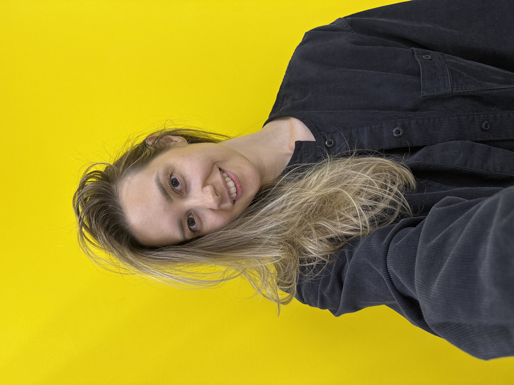 A cheerful young woman is featured in a diagonally oriented close-up against a vibrant, plain yellow background. She is smiling broadly, revealing her upper teeth, and looking directly into the camera with warm brown eyes. Her long, wavy, light blonde or brown hair frames her face and cascades towards the lower right of the frame. She wears a dark, possibly charcoal grey or black, corduroy shirt or jacket, identifiable by its distinct ridged texture and visible buttons on the placket and pockets. The lighting is bright and even, highlighting her features and the texture of her clothing. No specific time of day, weather, or location cues are visible beyond the plain studio-like background. There is no visible text. The overall scene suggests a posed portrait or selfie.