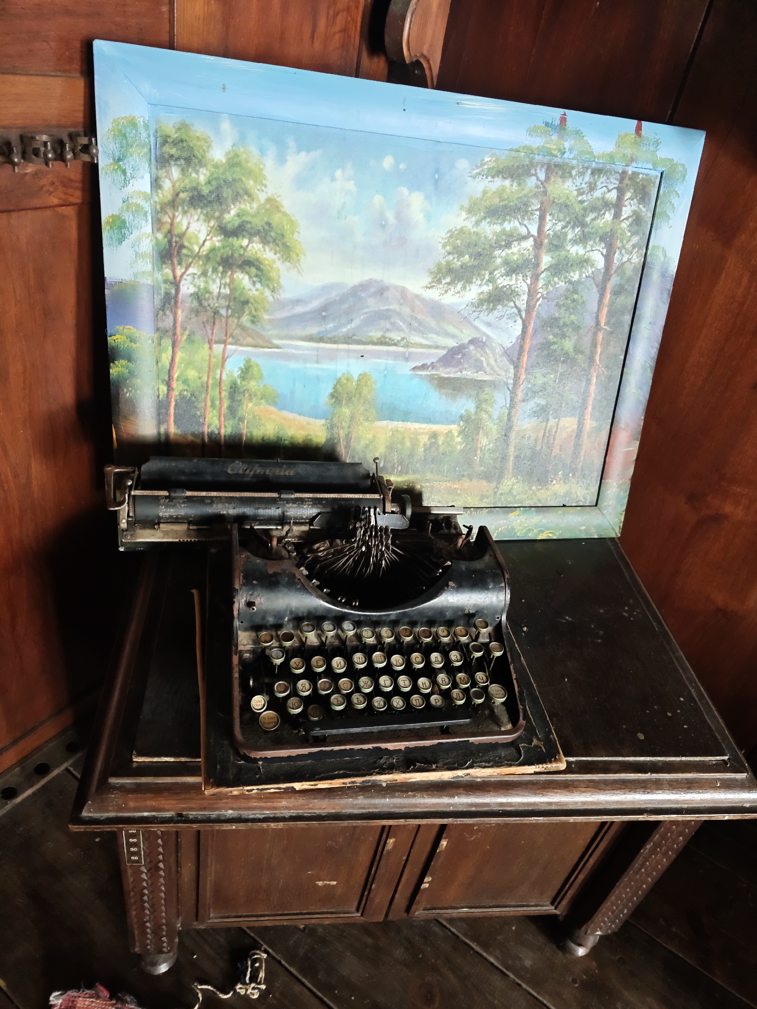 An antique Olympia typewriter rests on a dark wooden table, positioned in front of a framed landscape painting. The painting depicts a serene scene with a body of water, mountains in the distance, and tall coniferous trees in the foreground. The typewriter itself is black with visible metal parts and a full set of keys, some of which have Cyrillic lettering. The wooden table has ornate carvings on its legs and appears to be part of a larger piece of furniture, possibly a desk or cabinet. The background consists of dark wooden panels, suggesting an interior setting. The overall impression is one of a vintage or historical display, possibly within a home or museum. The location is identified as Lyutibrod, Bulgaria.