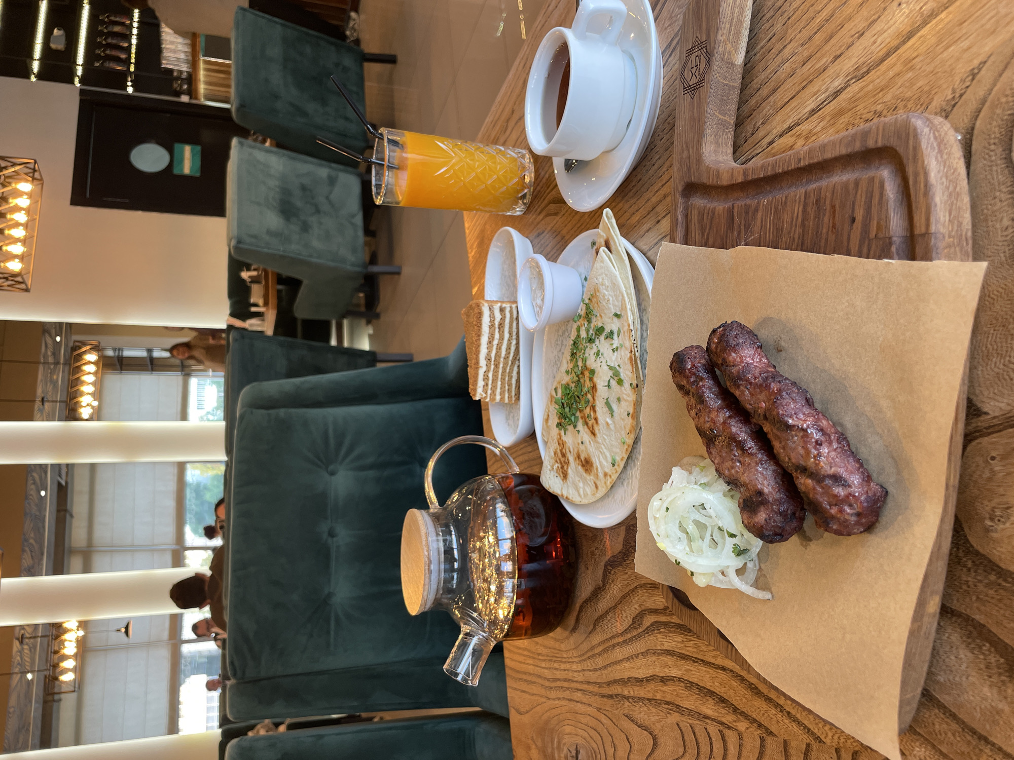 An overhead shot of a wooden table in a restaurant shows a meal and drinks. The meal consists of two grilled sausages, a flatbread topped with herbs, sliced onions, and a piece of cake. A glass of orange juice and a cup of tea are also on the table, alongside a glass teapot filled with tea. In the background, plush green chairs are visible, and behind them, large windows look out onto a street scene. Several people are seated at tables in the distance, and other diners can be seen in the background. The restaurant appears to be modern and well-lit, with contemporary light fixtures.