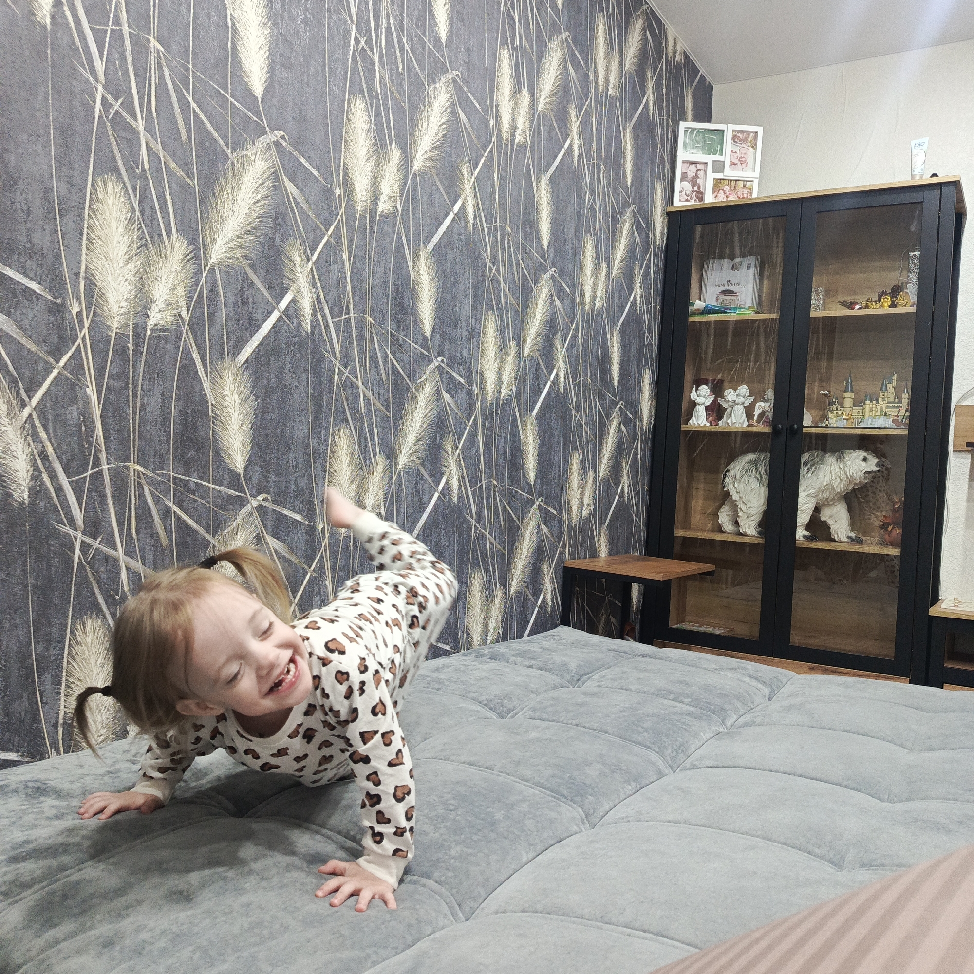 The image shows a young child playing on a light gray quilted sofa. The child, with blonde hair styled in two ponytails, is wearing a spotted pajama set and is in a crawling position, with a leg raised in the air. The child is smiling widely. Behind the sofa is a wall with a dark gray wallpaper featuring a light gold wheat-like pattern. To the right of the sofa is a dark-framed glass cabinet displaying various items, including figurines and a polar bear statue. Next to the cabinet is a small wooden table. The location is Марковский, Russia.