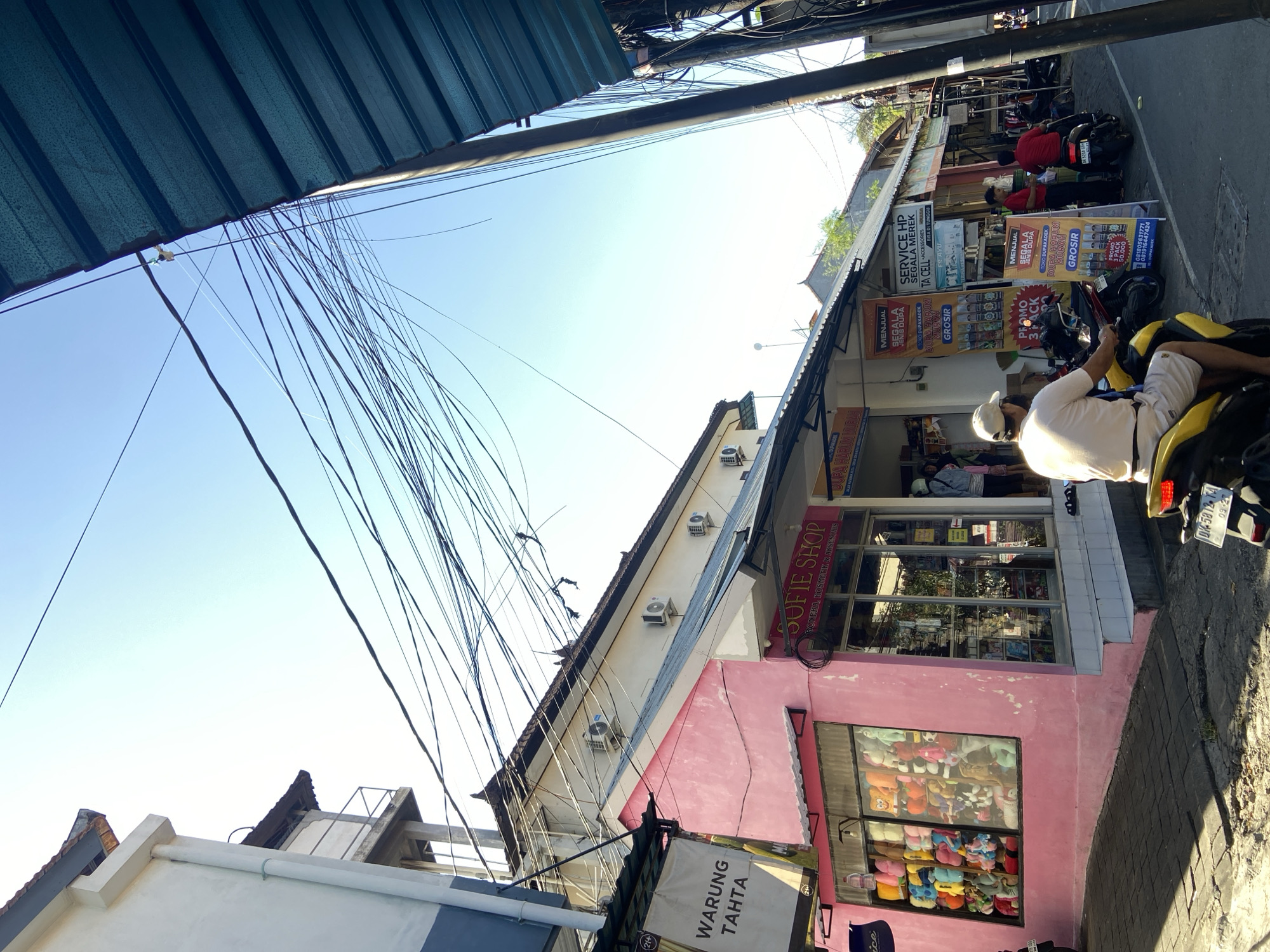 The image, taken at street level in Kuta, Indonesia, captures a sunny, bustling street scene. Buildings line the street, adorned with numerous overhead electrical wires. The foreground features a building painted pink, with a sign reading "SOFIE SHOP" and "WARUNG TAHTA". A window display showcases a collection of stuffed animals. A man wearing a white shirt and hat sits on a parked yellow scooter. Nearby, another scooter is parked, and in the background, several people are seen walking and riding scooters. A sign advertises cell phone repair services and the sale of incense.