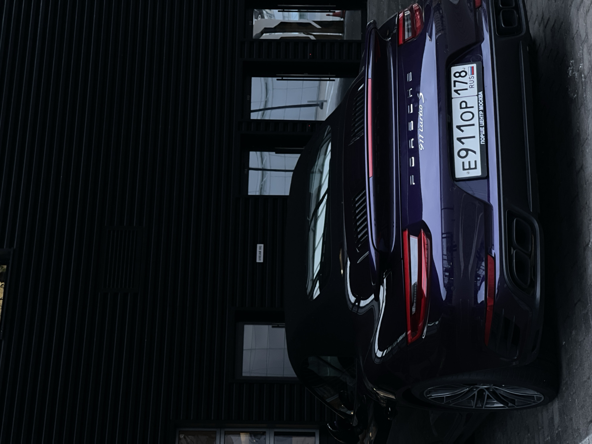 A dark purple Porsche 911 Turbo S convertible is parked in front of a building with a dark, horizontally slatted facade. The car's convertible top is up. The rear of the car is visible, showing its taillights, spoiler, and quad exhaust pipes. A license plate with the characters "E911OP 178" and "RUS" is mounted on the rear bumper. Below the license plate, text reads "ПОРШЕ ЦЕНТР МОСКВА" which translates to "Porsche Center Moscow." The ground appears to be paved with grey cobblestones. The building features glass doors with dark frames.