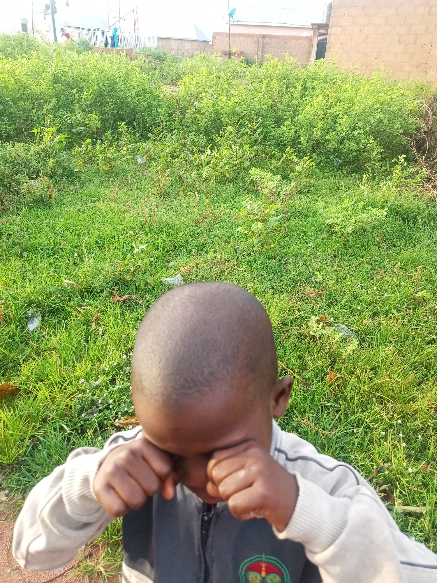 A young boy, likely under ten years old, stands in a grassy, overgrown area with his face obscured by his hands as he appears to be crying. He is wearing a light-colored hooded sweatshirt with dark accents and a dark zippered jacket underneath. The background consists of a dense thicket of green foliage, with some scattered trash visible on the ground. In the distance, several buildings and structures are partially visible, suggesting an urban or semi-urban environment. The lighting indicates it is daytime. The scene is located in Tempure, Nigeria.