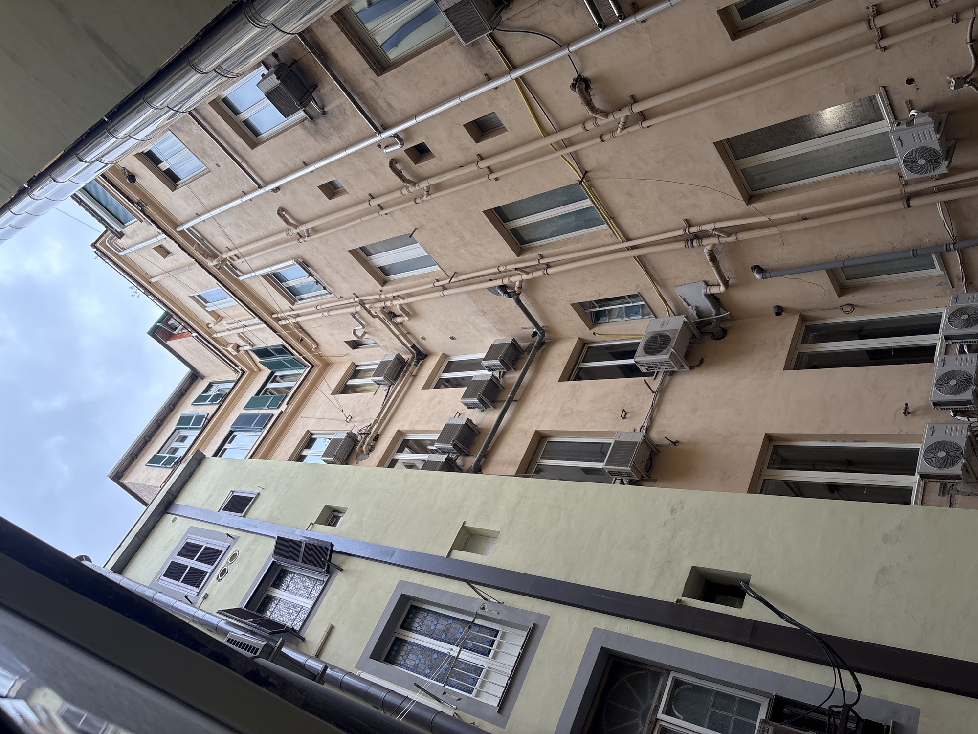 The image shows an upward-angle view of an urban courtyard in Rome, Italy. Two buildings, one painted a pale yellow and the other a light peach, face each other, creating a narrow, enclosed space. The buildings are multi-story, with numerous windows visible on their facades. Many of the windows have open or closed shutters, some are adorned with decorative glass or small balconies.
Several air conditioning units are mounted on the exterior walls of both buildings, indicating a warm climate. A complex network of pipes, likely for plumbing and drainage, runs vertically and horizontally along the peach-colored building. Electrical conduits and wiring are also visible. The sky above is overcast with a muted, greyish hue. The perspective is distorted due to the upward angle, emphasizing the height and claustrophobic nature of the courtyard. There are no people or animals visible in the scene. The overall impression is one of a densely built urban environment with functional elements integrated into the architecture.