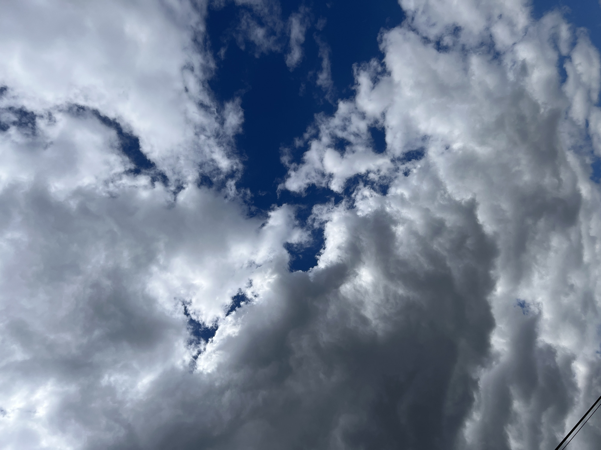 The image depicts a sky with scattered clouds over the city of Sheptytskyi, Ukraine. The clouds are varied in density and color, ranging from bright white to dark gray, suggesting a partly cloudy or approaching stormy condition. Patches of deep blue sky are visible between the clouds. A thin, dark diagonal line, possibly a power line, is visible in the bottom left corner of the frame. The perspective is upward, looking towards the sky.