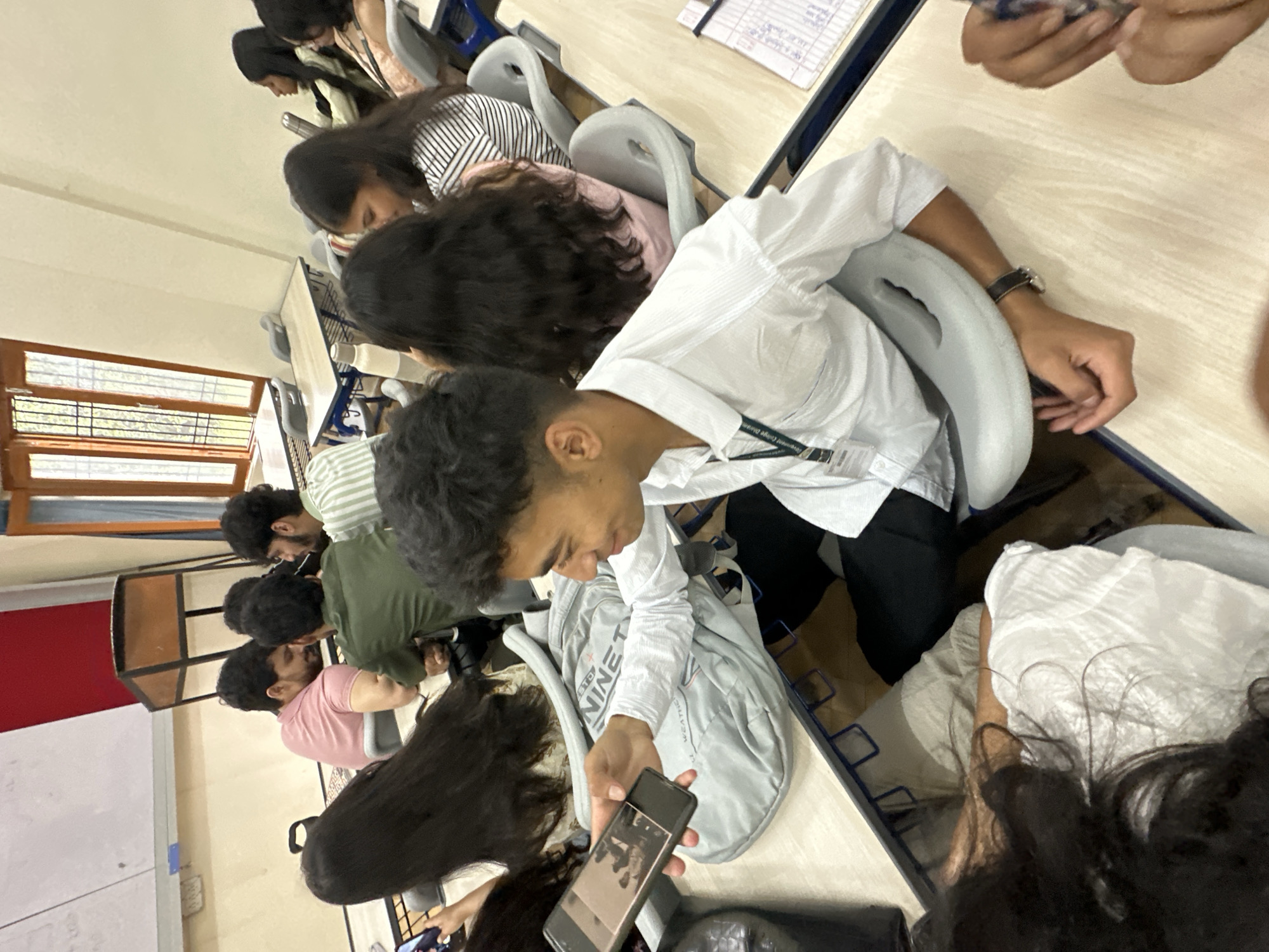 The image depicts a classroom or lecture hall setting where a group of young adults are seated at desks. In the foreground, a young man in a white shirt is holding up a smartphone, appearing to show something on the screen to someone off-camera. He is wearing a lanyard with an ID badge. Beside him, a light grey backpack with text is visible on the desk. Several other individuals are seated around him, some facing away from the camera, others partially visible. The room has rows of desks with blue frames and light-colored tabletops. In the background, there are windows with wooden frames and a whiteboard or projection screen on the wall. The overall atmosphere suggests a casual educational environment.